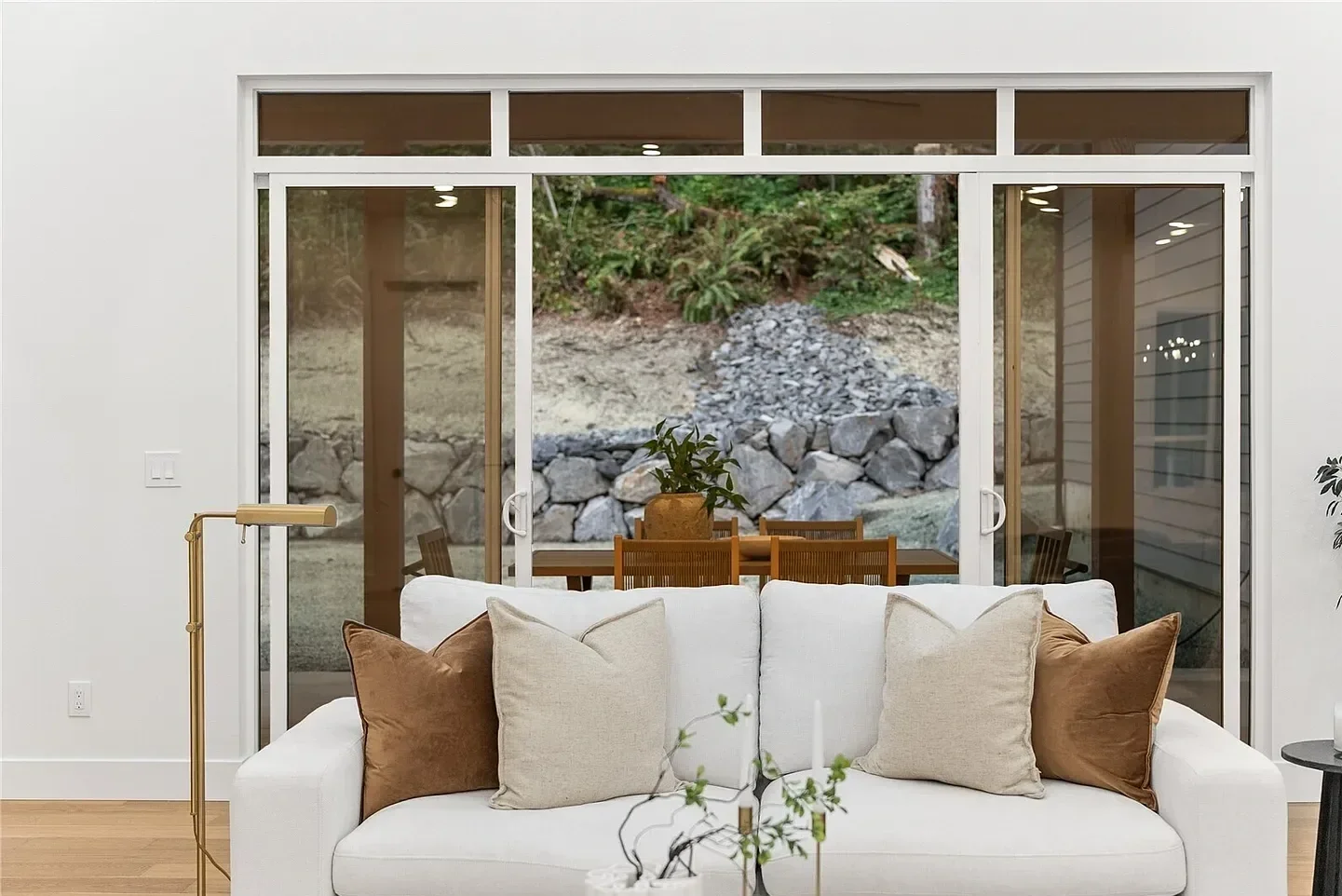 Living room with a white sofa, beige and brown pillows, a gold floor lamp, and a sliding glass door leading to an outdoor patio with a wooden dining table, chairs, and a potted plant, overlooking a rocky hillside with greenery.