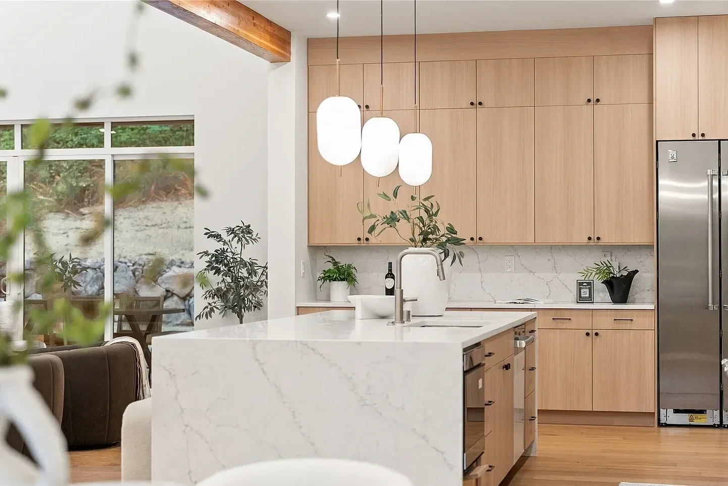 Modern kitchen with wooden cabinets, white marble countertops, pendant lights, potted plants, stainless steel refrigerator, and large sliding glass door leading outside.