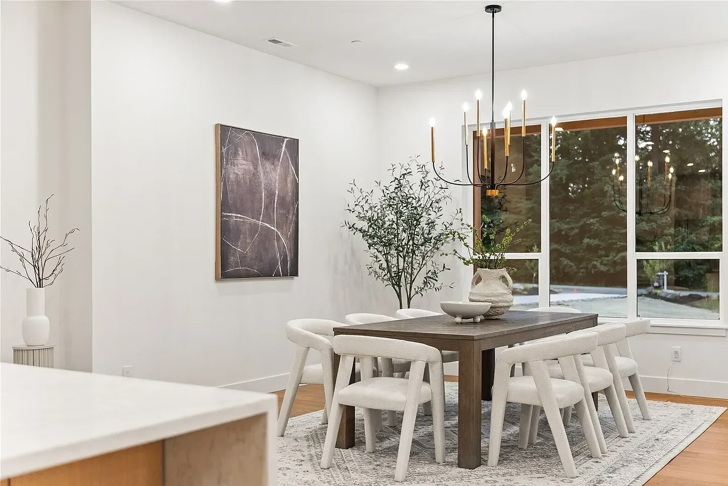 Modern dining room with a wooden table, eight white chairs, a chandelier, large windows, artwork on the wall, and decorative plants.
