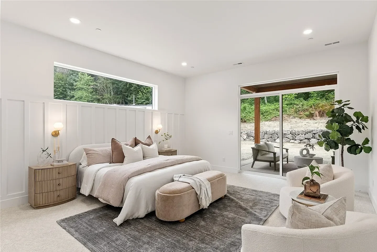Bright bedroom with white paneled wall behind bed, bedside tables, and sliding glass door leading to outdoor patio with chairs and a tree.