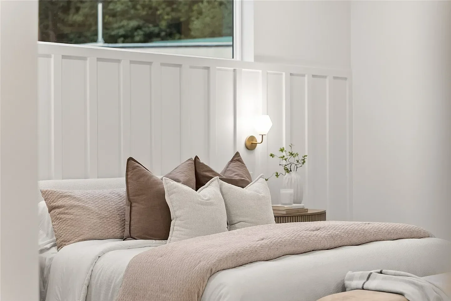 A cozy bedroom with a bed decorated with beige and white pillows, a beige blanket, a wooden nightstand with a potted plant, a modern wall sconce, and a window overlooking greenery.