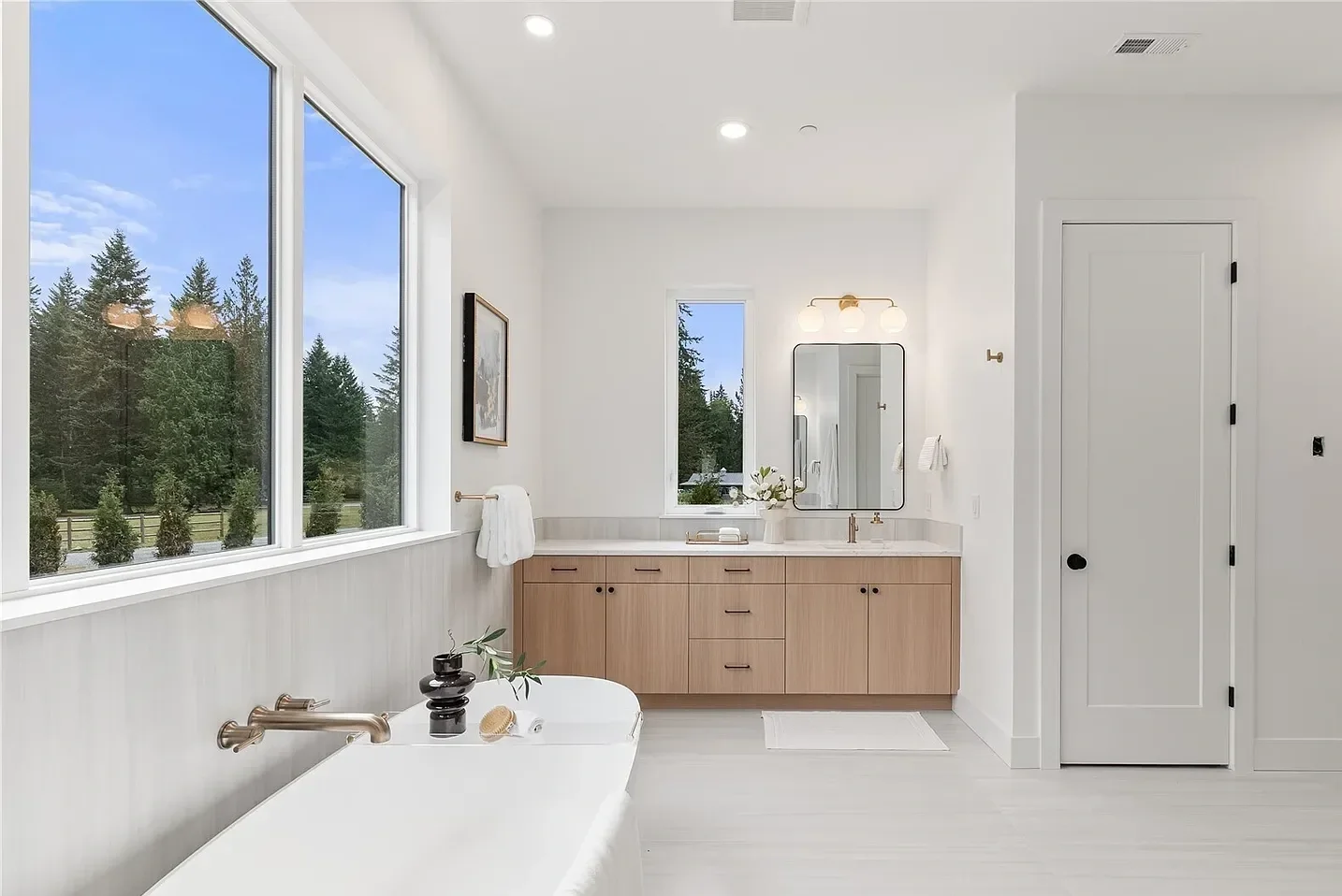 Modern bathroom with large window showing outdoor trees, white walls, wooden vanity, standing mirror, and bathtub with decorative accessories.