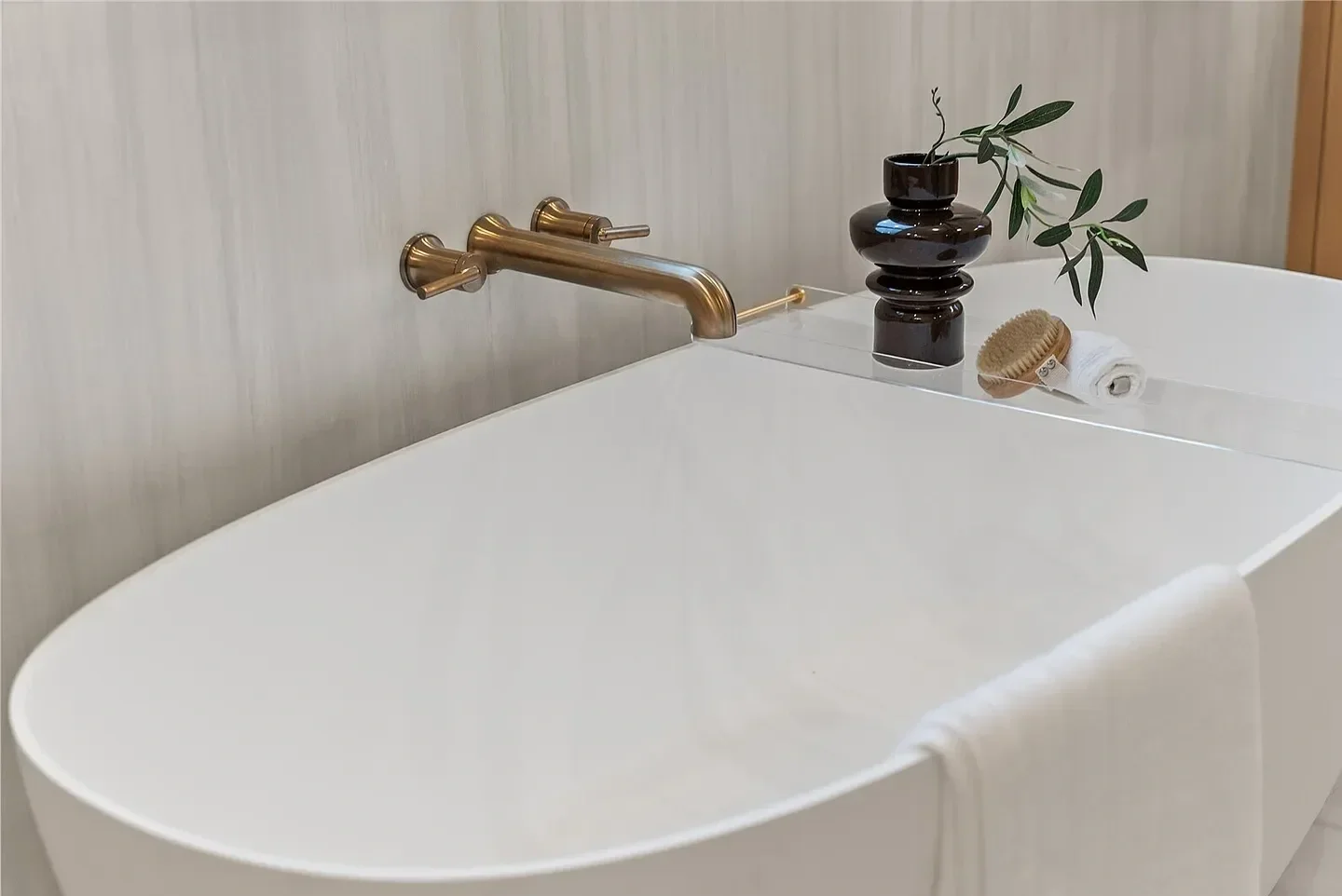 A modern bathroom with a white bathtub, a golden wall-mounted faucet, a black vase with greenery, a bath brush, and a rolled white towel.
