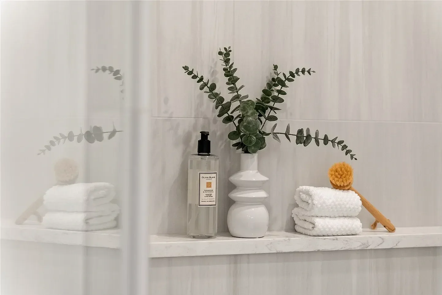 Bathroom shelf with a clear soap dispenser, a white ceramic decorative vase with green foliage, three rolled white towels, and a wooden body brush with a round orange loofah on top.