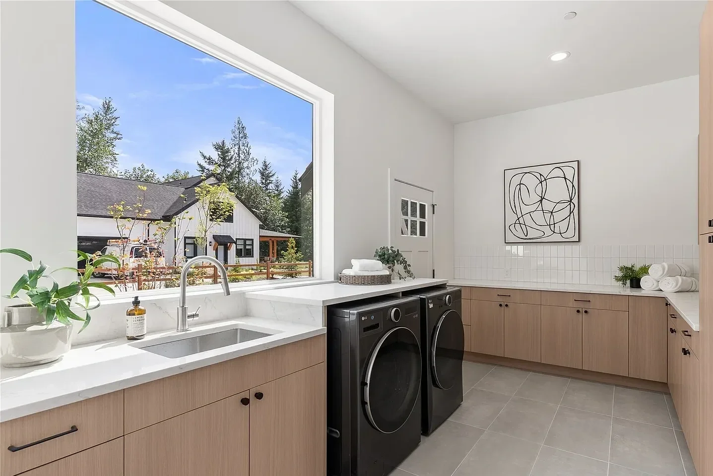 A modern laundry room with a large window showing an outside view of trees and houses. The room has light wood cabinets, a white marble countertop, a black front-loading washer and dryer, and minimalistic decor including a potted plant, towels, and a