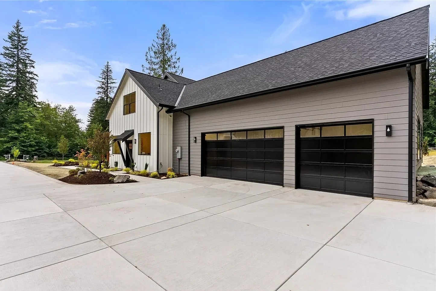 Modern house with black garage doors, gray siding, and large windows, surrounded by landscaped yard and trees.