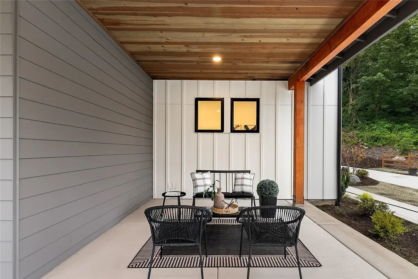 Outdoor covered patio with black chairs, a small table, and decorative pillows, enclosed by a white wall and a gray wall, with a wooden ceiling and visible outdoor greenery.