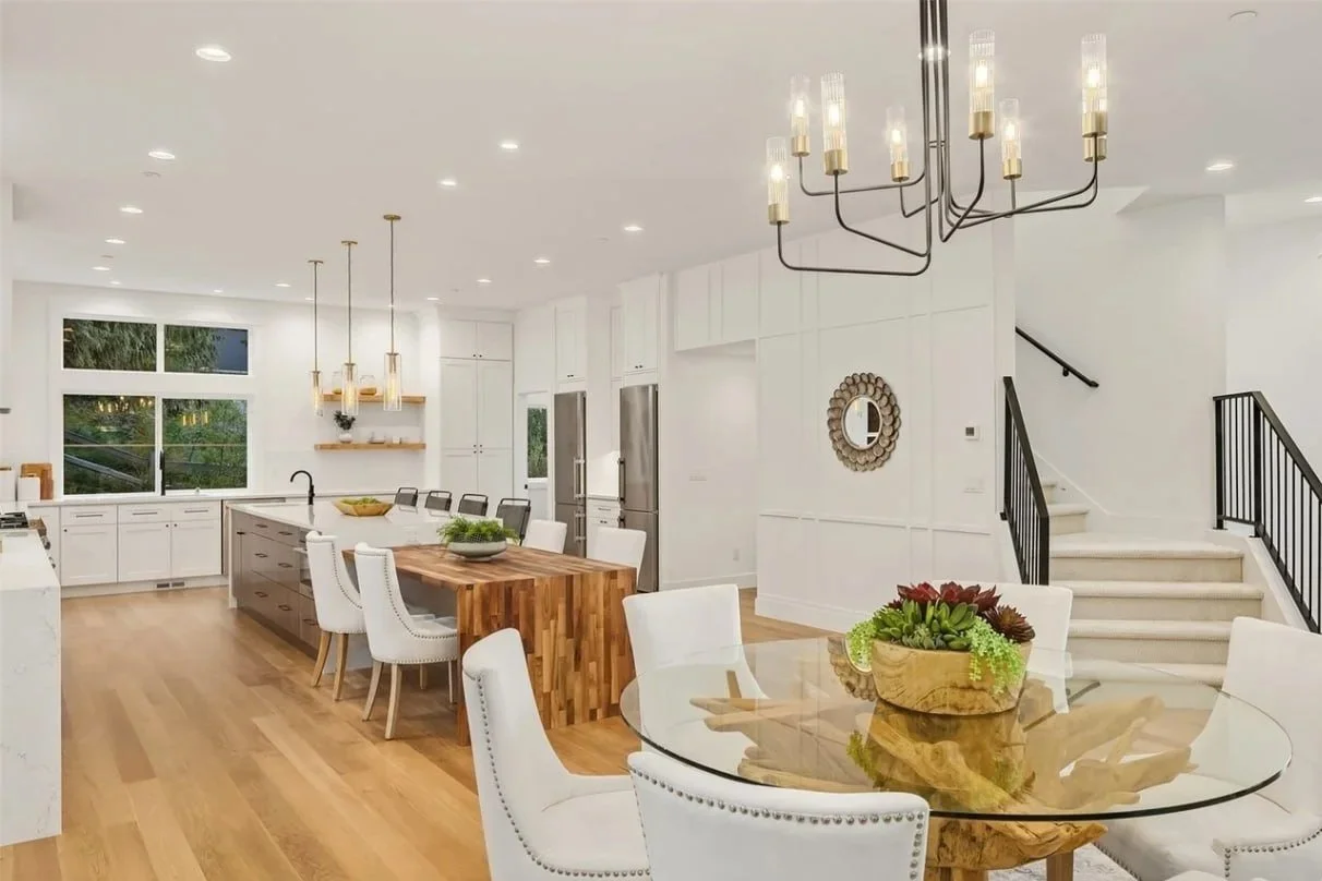 Open kitchen and dining area with white cabinets, a wooden kitchen island, and a glass-top dining table with white chairs. Modern lighting fixtures and large windows.