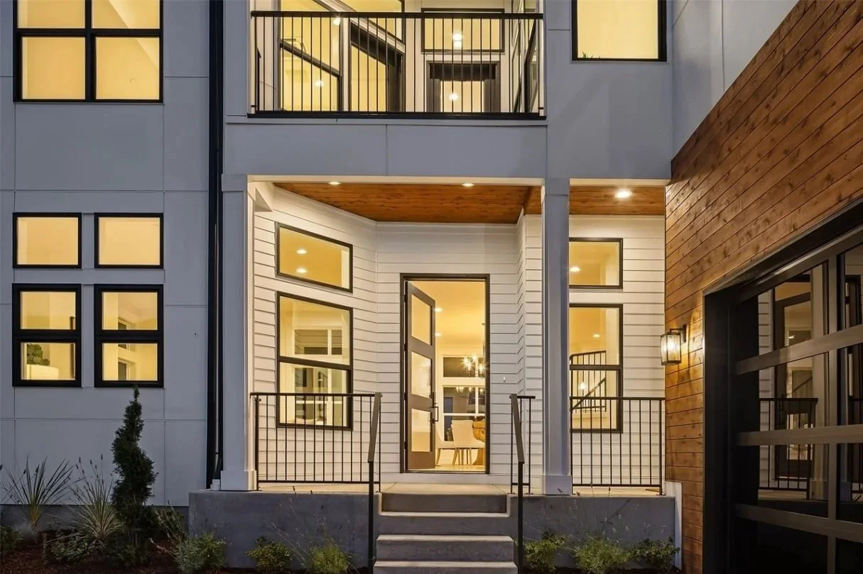 Modern multi-story house with white and wooden exterior, illuminated windows, front porch, black railings, and small landscaped garden.
