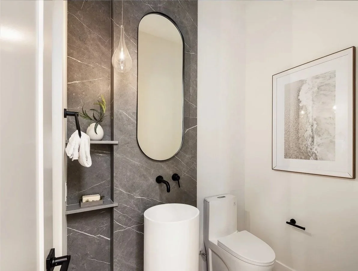 Modern bathroom with gray marble wall, oval mirror, white cylindrical sink, and toilet, decorated with framed artwork, a shelved caddy with a plant and towels, and a pendant light.