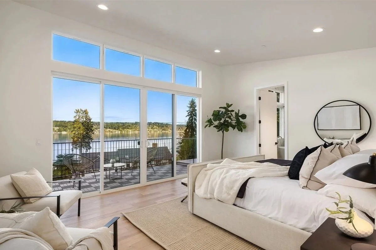 Bright, modern bedroom with a large sliding glass door leading to a balcony that overlooks a lake and trees. The room has white walls, a white bed with multiple pillows, a small black round mirror, a potted plant, and minimal decor.