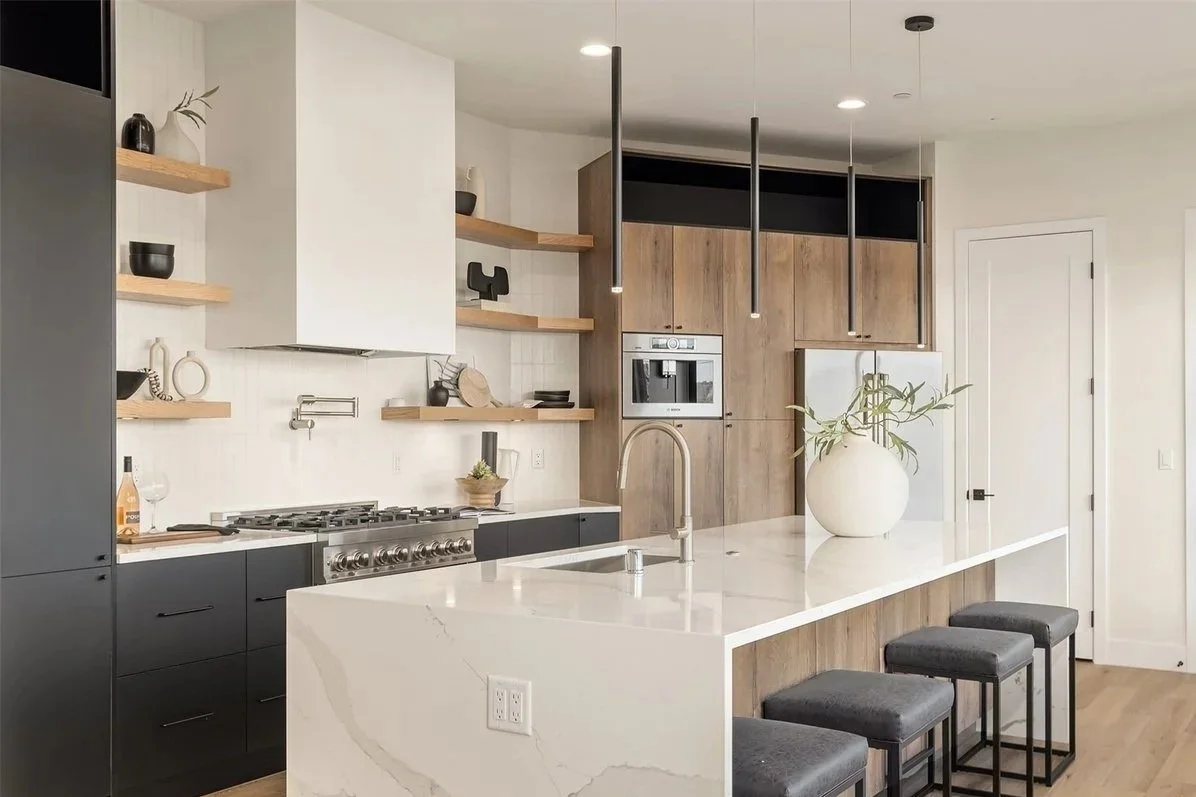 Modern kitchen with white marble island, black lower cabinets, wooden open shelves, and light wood upper cabinets. Decorative vases and bowls are on the shelves, with a large white vase with green plants on the island. There are three gray barstools 