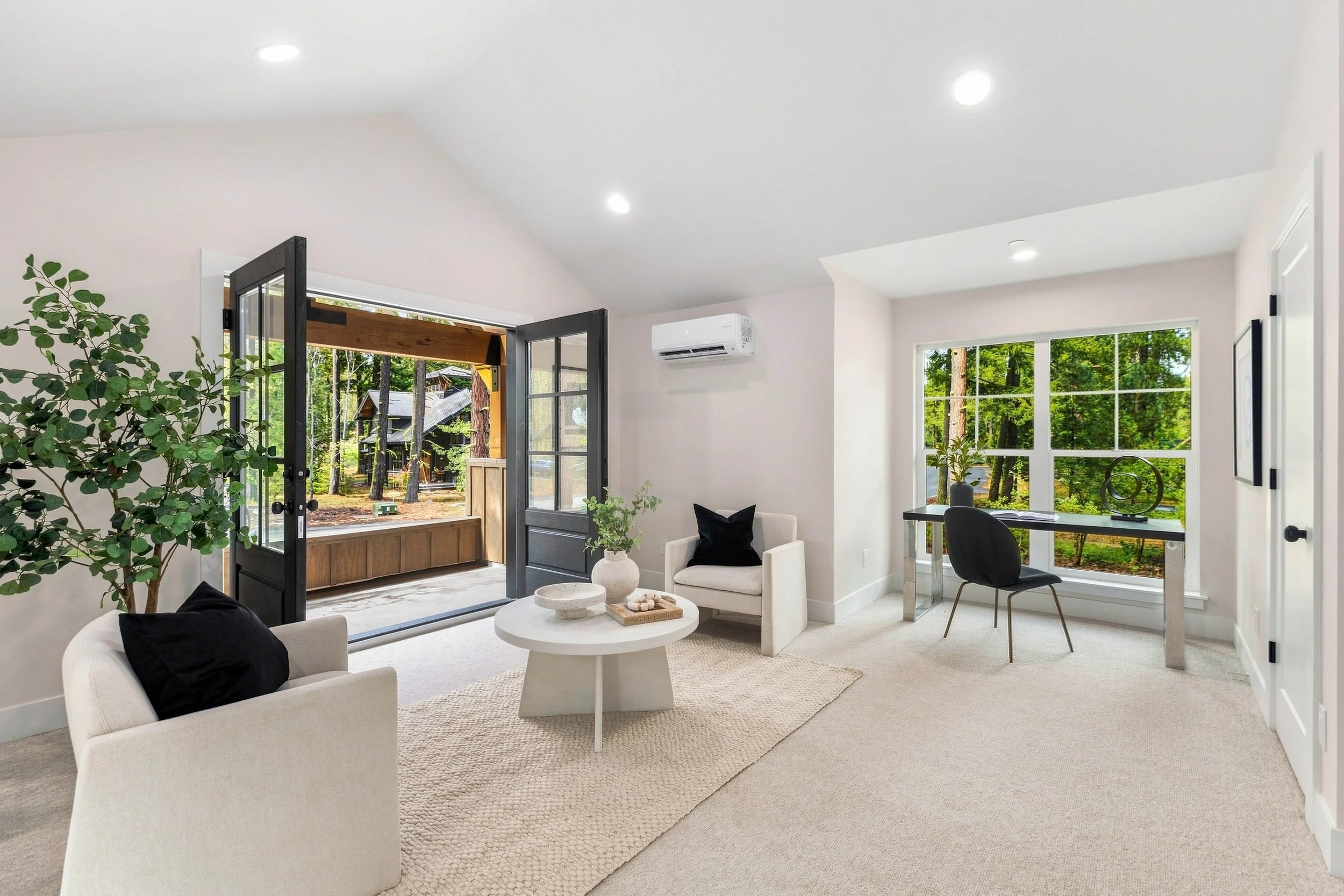 Bright living room with large windows looking onto a wooded yard, featuring white walls, a white couch with black pillows, a round white coffee table, a plant, and a workspace with a desk and black chair.