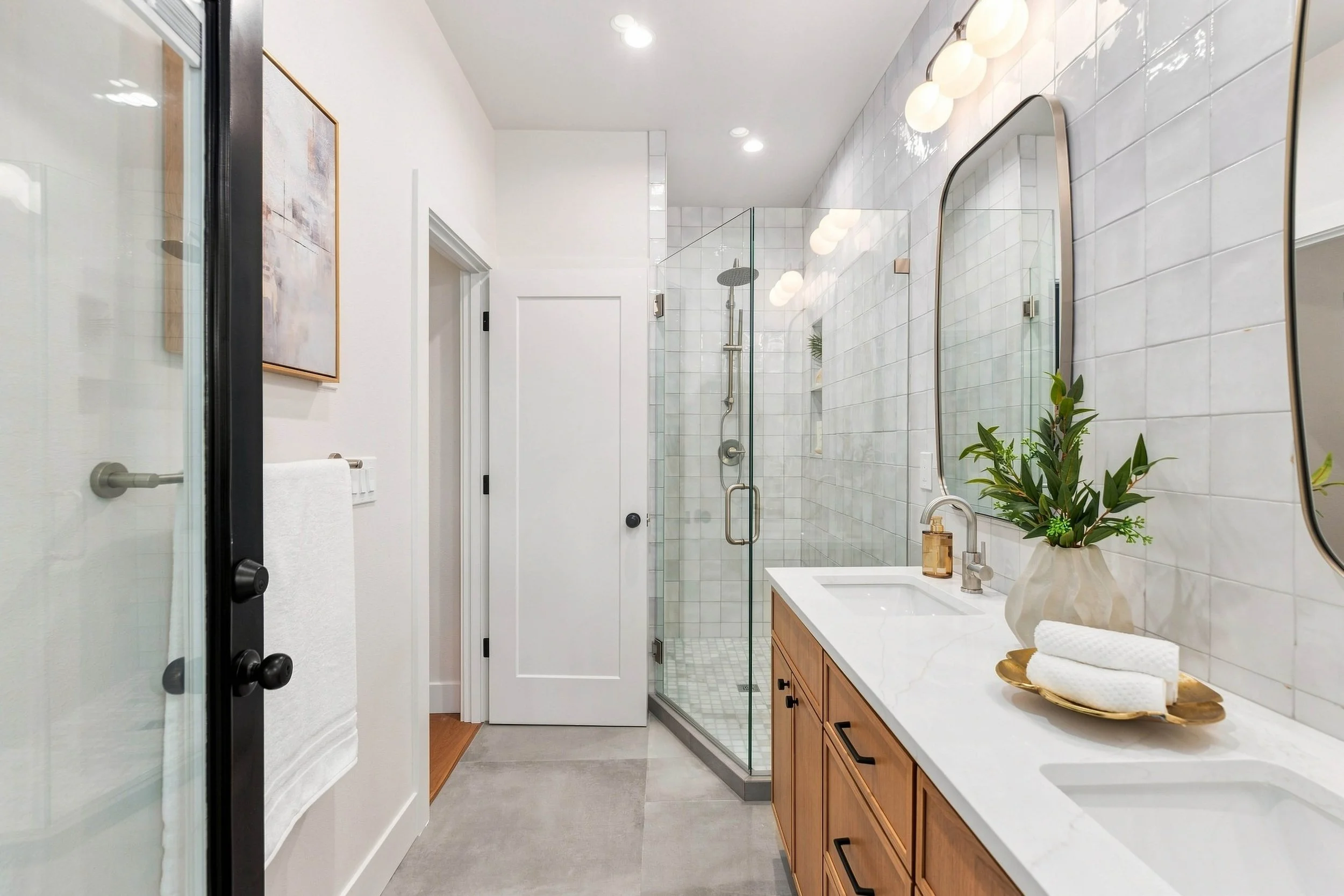 Modern bathroom with glass shower enclosure, white subway tile walls, wooden vanity, large mirror, and greenery in a vase.