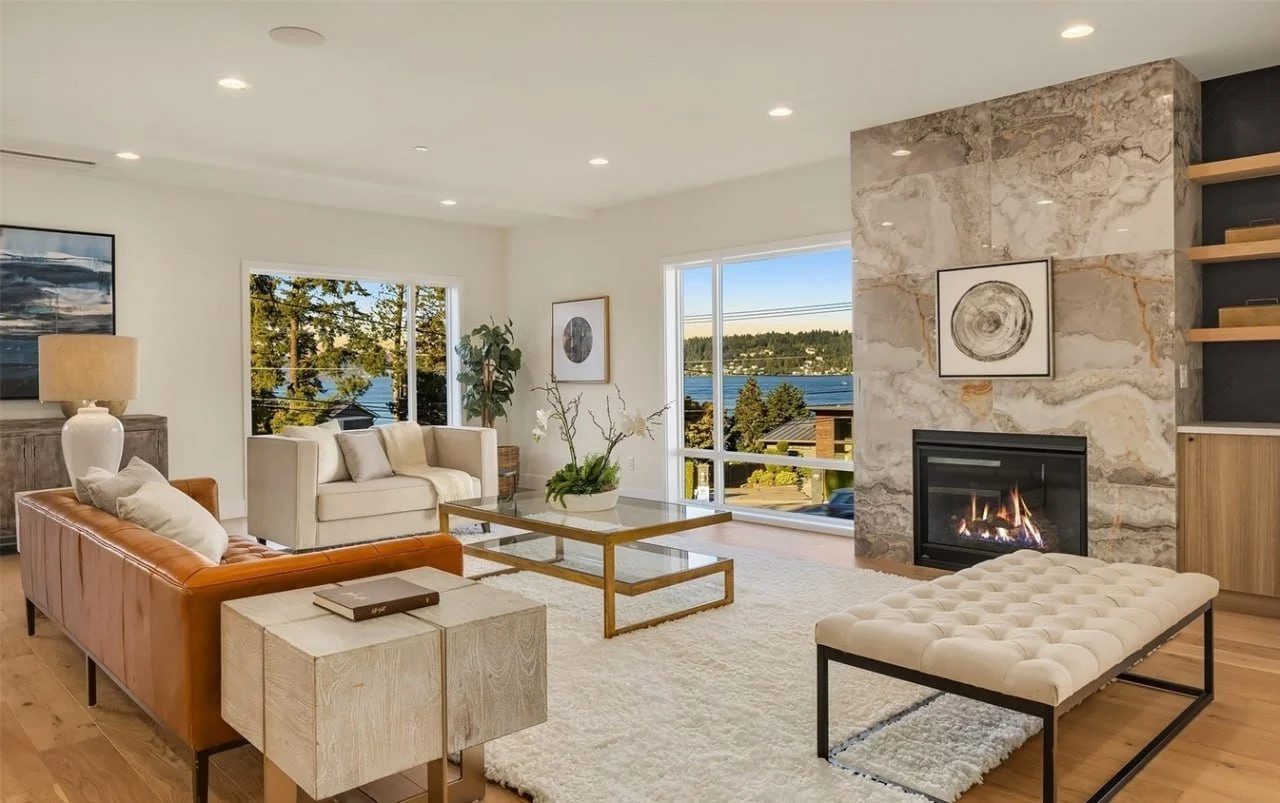 Living room with white and tan sofas, a glass coffee table with a plant, a fireplace with a marbled wall, large windows showing views of a lake and trees, wooden shelves, and a light-colored rug.