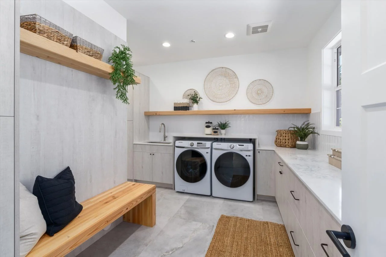 Laundry room with washer and dryer, wooden bench with a black pillow, white cabinetry, a marble countertop, wicker baskets, and decorative wall art.