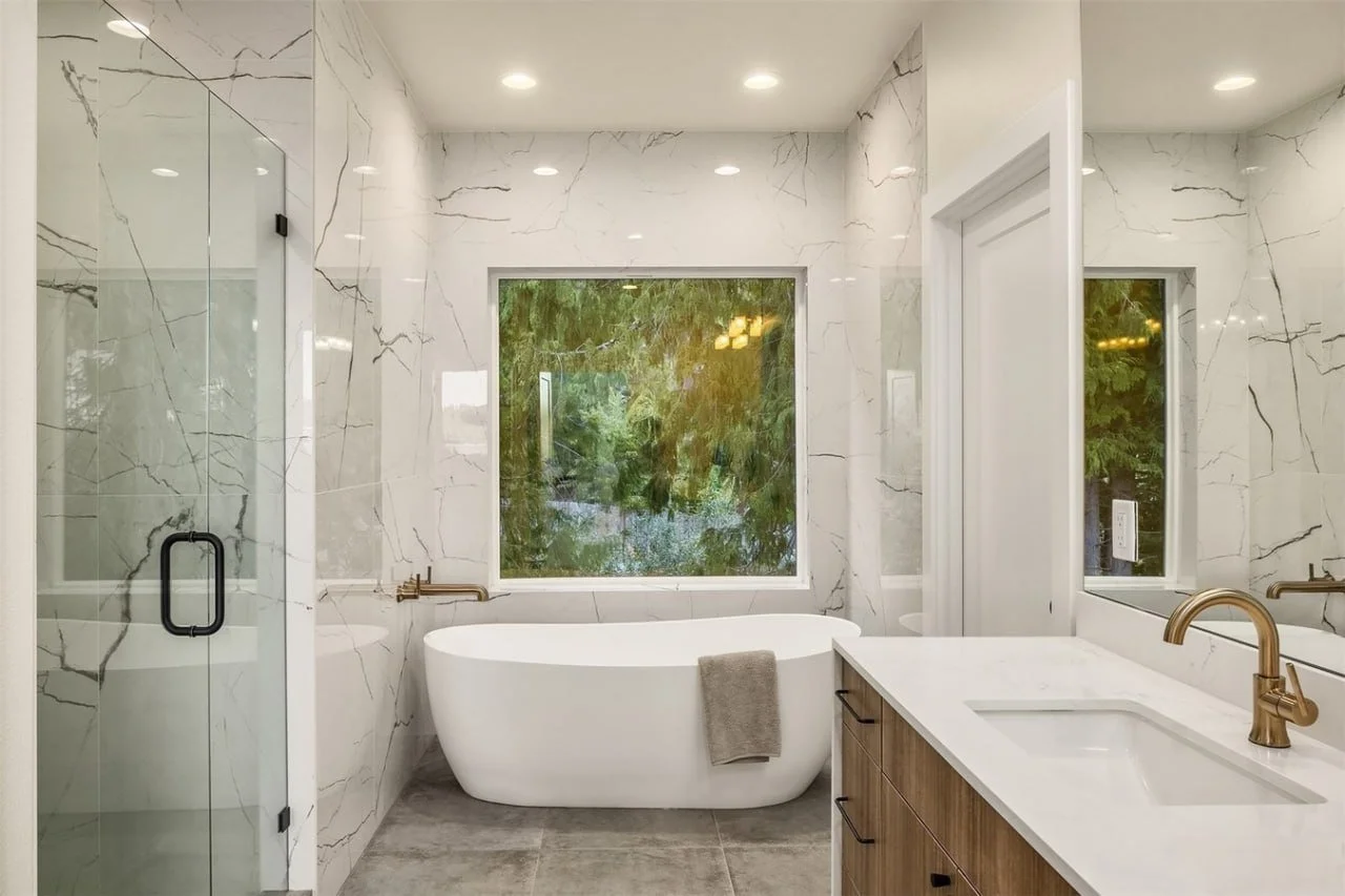 Modern bathroom with marble walls, a freestanding bathtub, a glass shower enclosure, a wooden vanity with a white countertop, and a large window showing greenery outside.