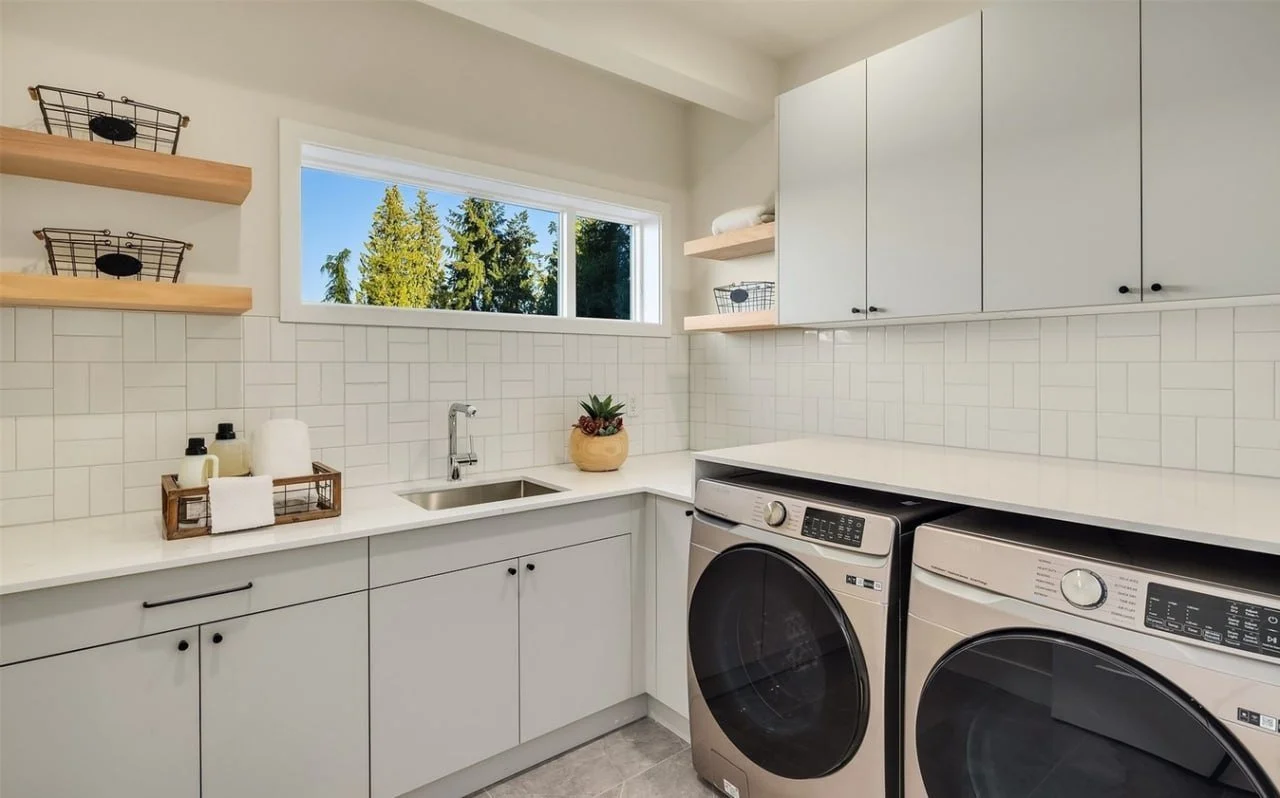 Laundry room with white cabinets, a laundry sink, a plant, and a window showing trees outside. There are open shelves with baskets and a front-loading washing machine and dryer.
