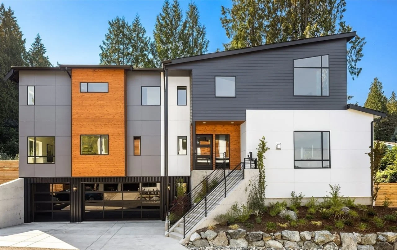 Modern multi-story house with gray, white, and wood-paneled exterior, large windows, front stairs, and landscaped yard.