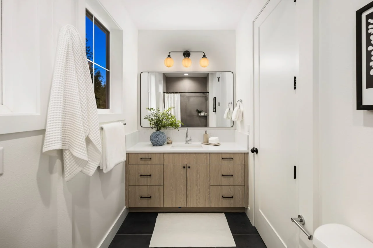 Modern bathroom with white walls, a wooden vanity, a large mirror, and a window showing a blue sky. Decor includes a plant in a blue pot, towels, and framed artwork.