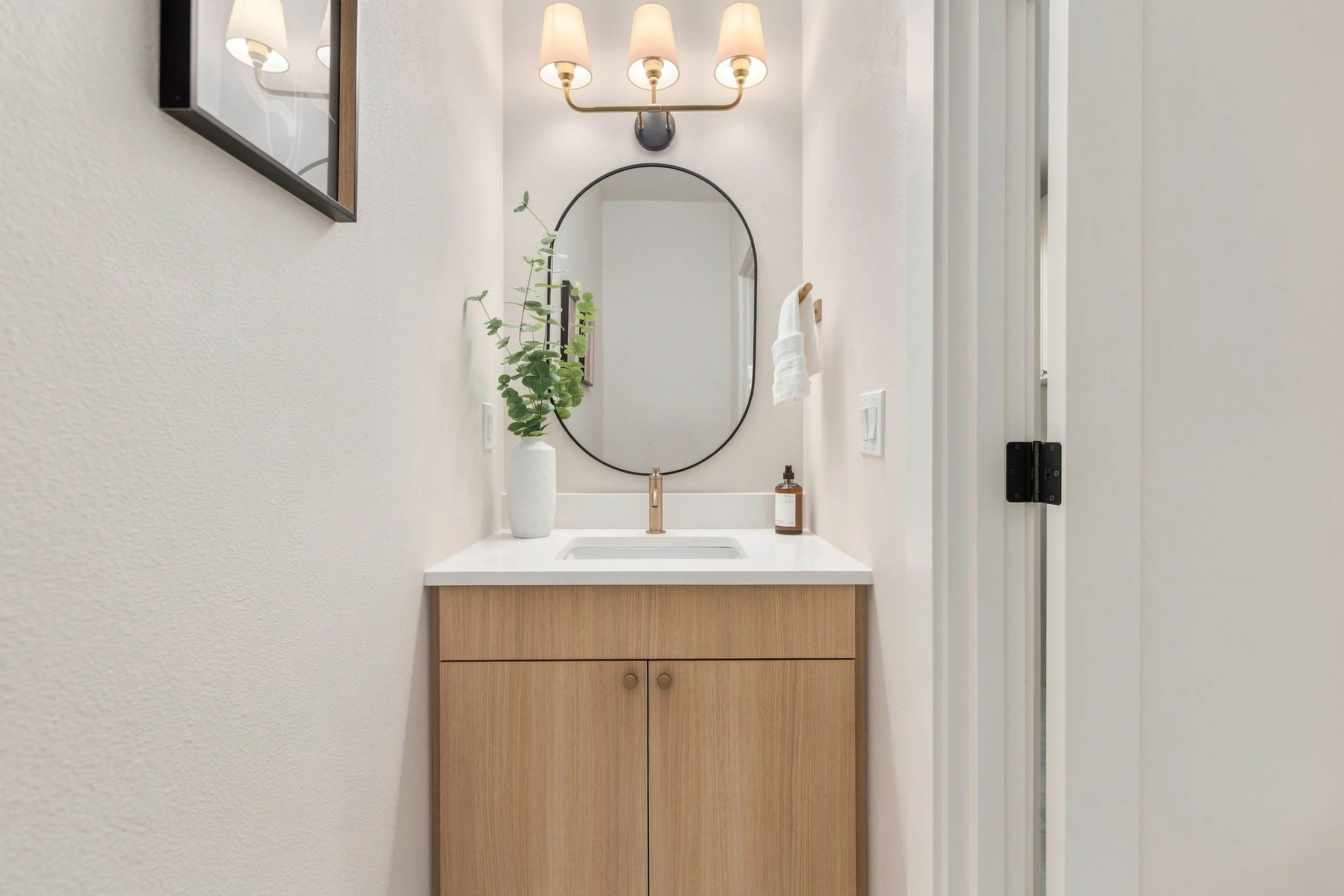 Bathroom vanity with a white countertop, a small sink, a gold faucet, a tall white vase with green branches, a wall-mounted mirror, and a four-light wall sconce above the mirror.