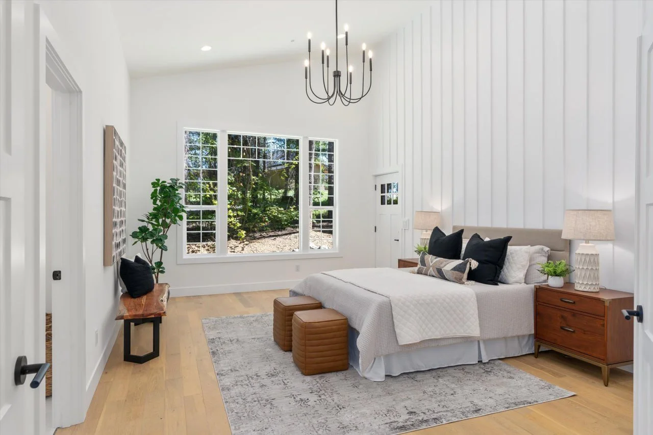 Bright bedroom with a large window overlooking greenery, a chandelier hanging from the ceiling, a bed with black and cream pillows, wooden nightstands with lamps, a wooden bench at the foot of the bed, and a potted plant near the window.