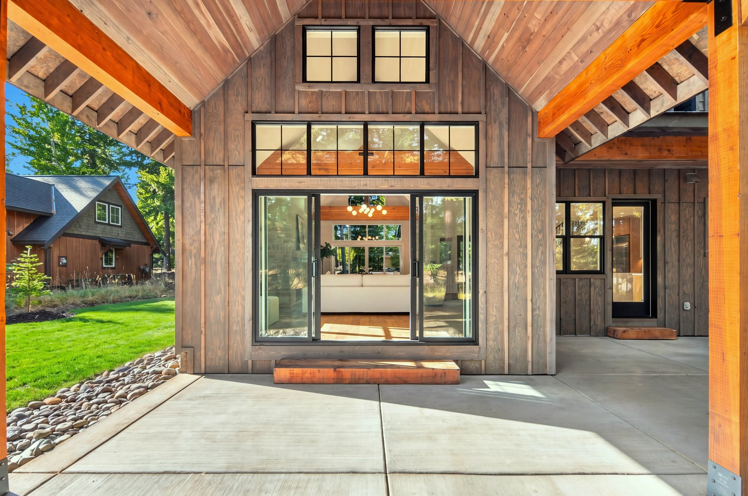 View of a modern house exterior with large glass doors and wooden siding, showing an open interior living space with a chandelier and patio in a green yard.