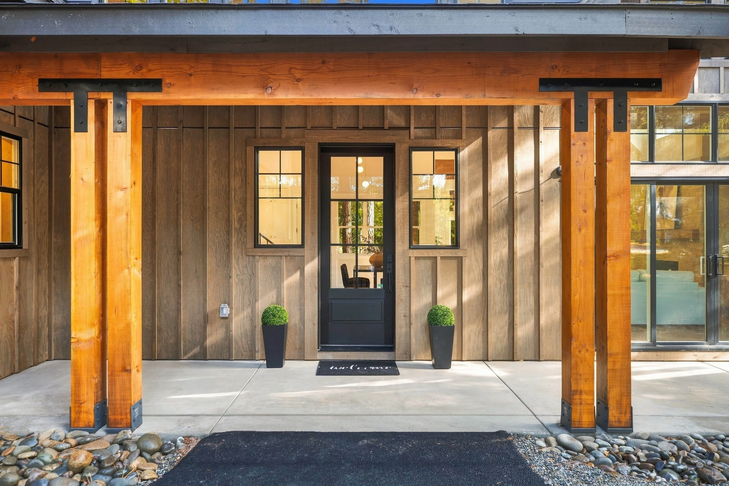 Modern house front with wooden and metal accents, large windows, and an entryway flanked by potted plants, featuring a welcoming doormat.