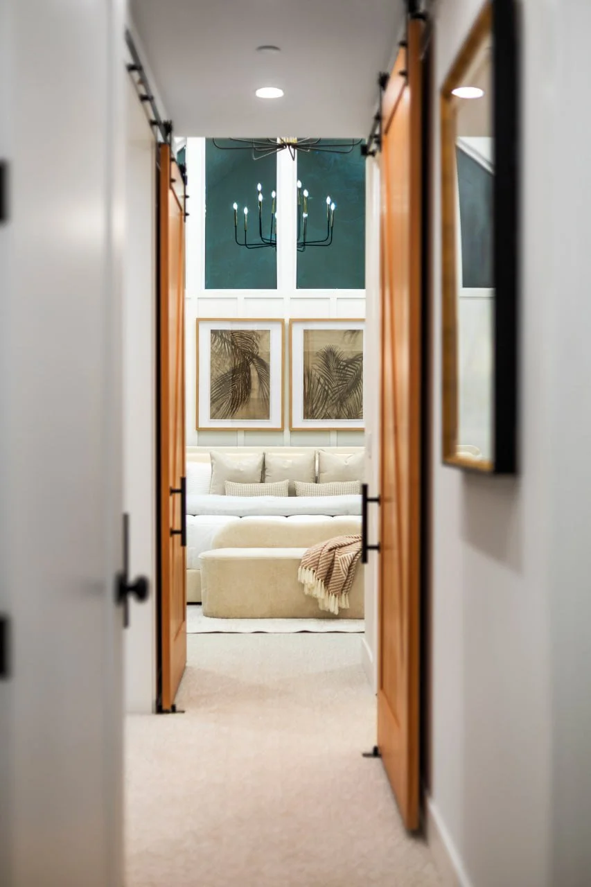 View through a narrow hallway into a living room with a beige sofa, framed artwork of palm leaves above, and a dark chandelier on the ceiling.