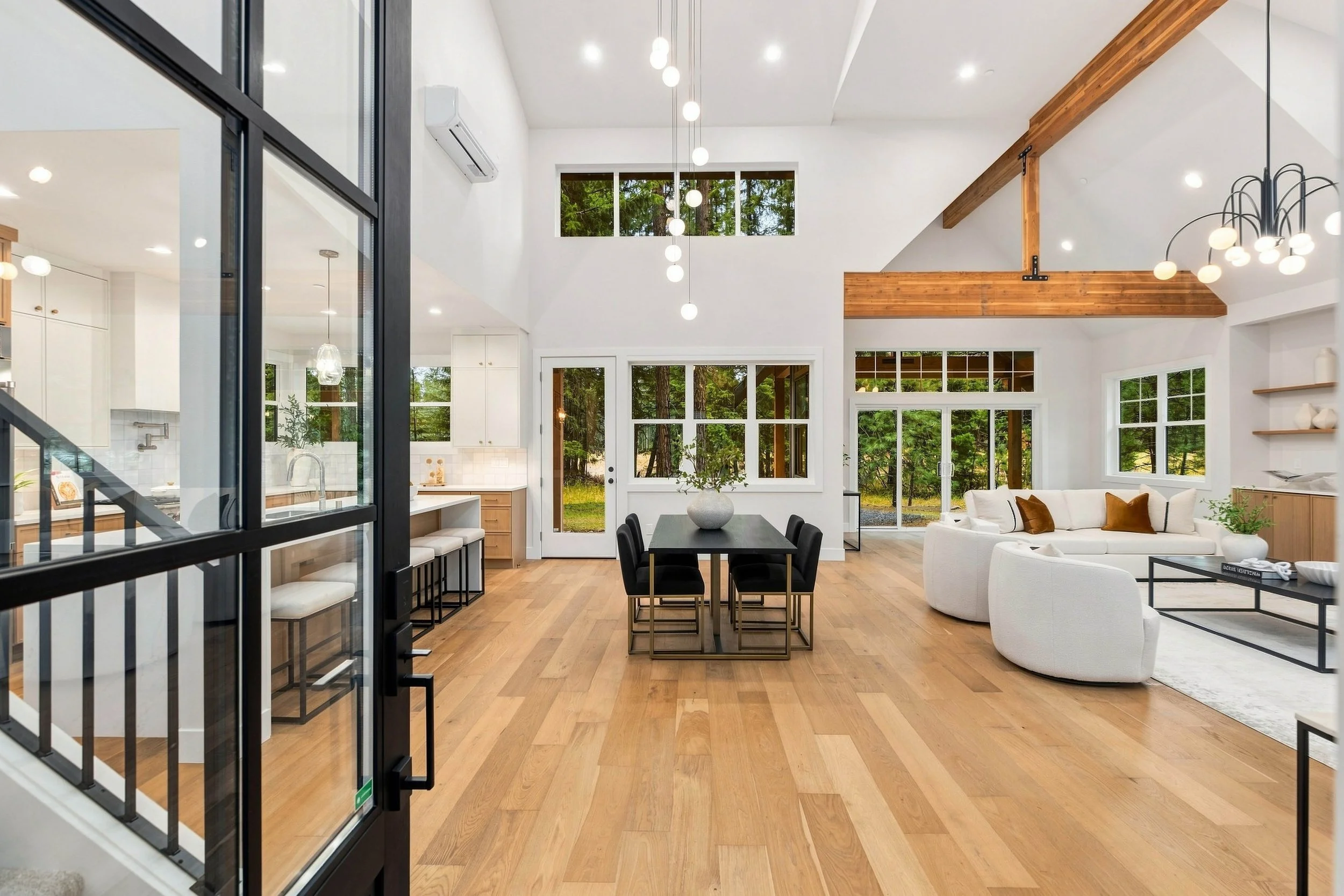 Bright, modern living room with high white ceiling, large windows showing trees outside, wooden beams, white walls, hanging spherical lights, a white sectional sofa with pillows, a black dining table with chairs, and wooden floors.