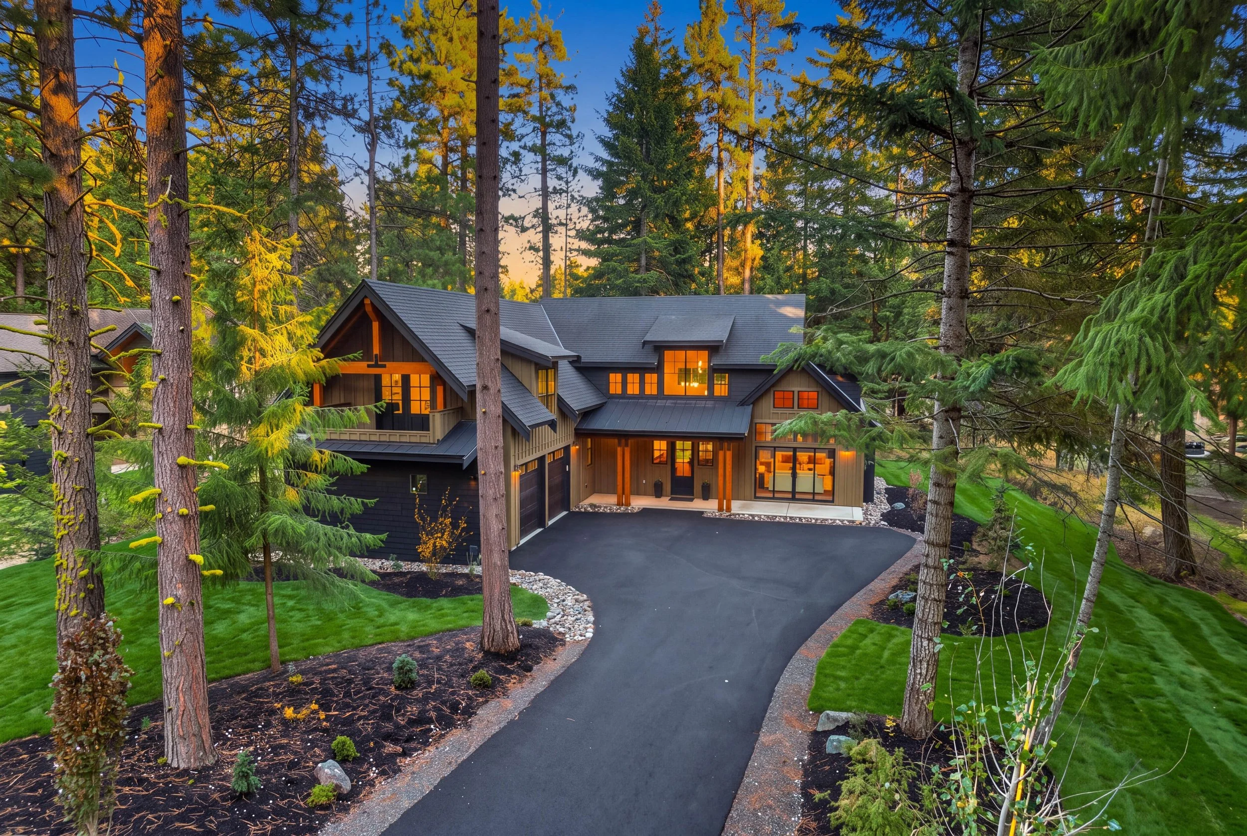 A modern house with dark exterior and large windows glowing with warm interior light, surrounded by tall pine trees and lush green grass in a wooded area during sunset.