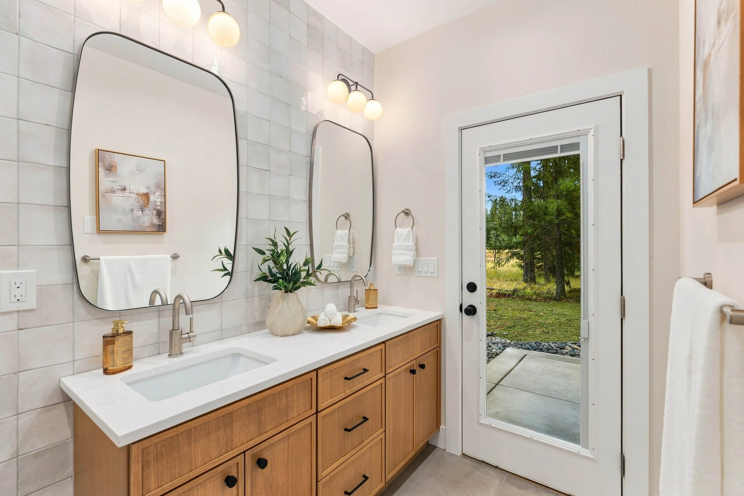Modern bathroom with double sinks, large mirrors, and white tiled wall. Natural light from a door with window showing trees outside, wood vanity, wall-mounted lights, and decorative items on countertop.
