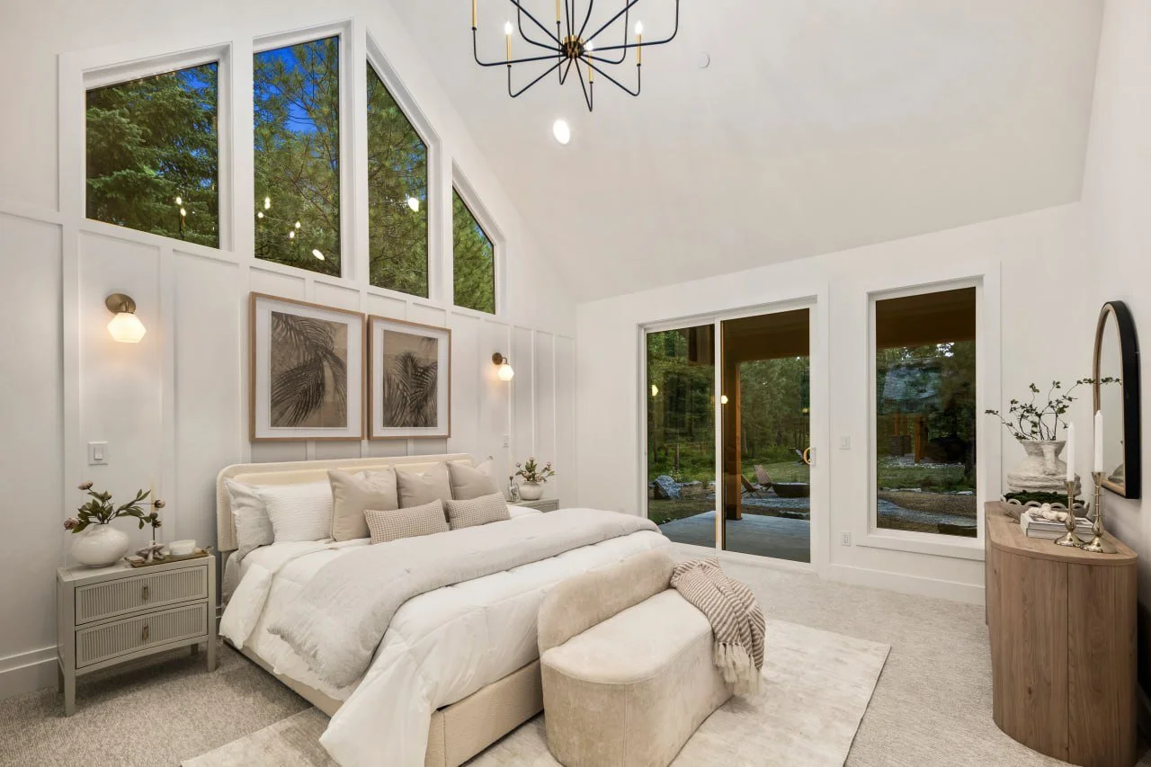 A bedroom with a high vaulted ceiling, large windows and glass sliding doors showing greenery outside. The room has a white bed with multiple pillows, nightstands with plants, and framed artwork above the bed. There is a light-colored bench at the fo
