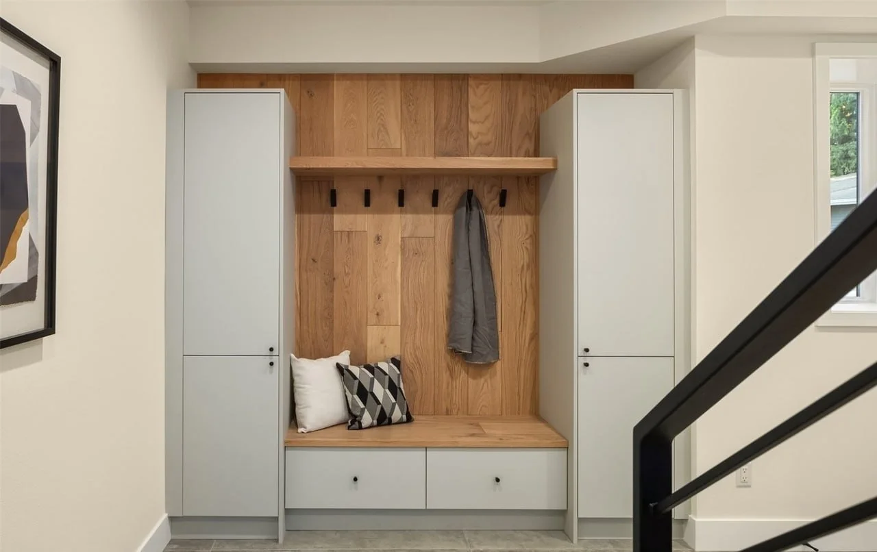 A modern entryway with white cabinets on each side, a wooden wall panel with black hooks, a bench with two pillows, and a gray jacket hanging on one of the hooks. There is a window to the right and a staircase railing in the foreground.