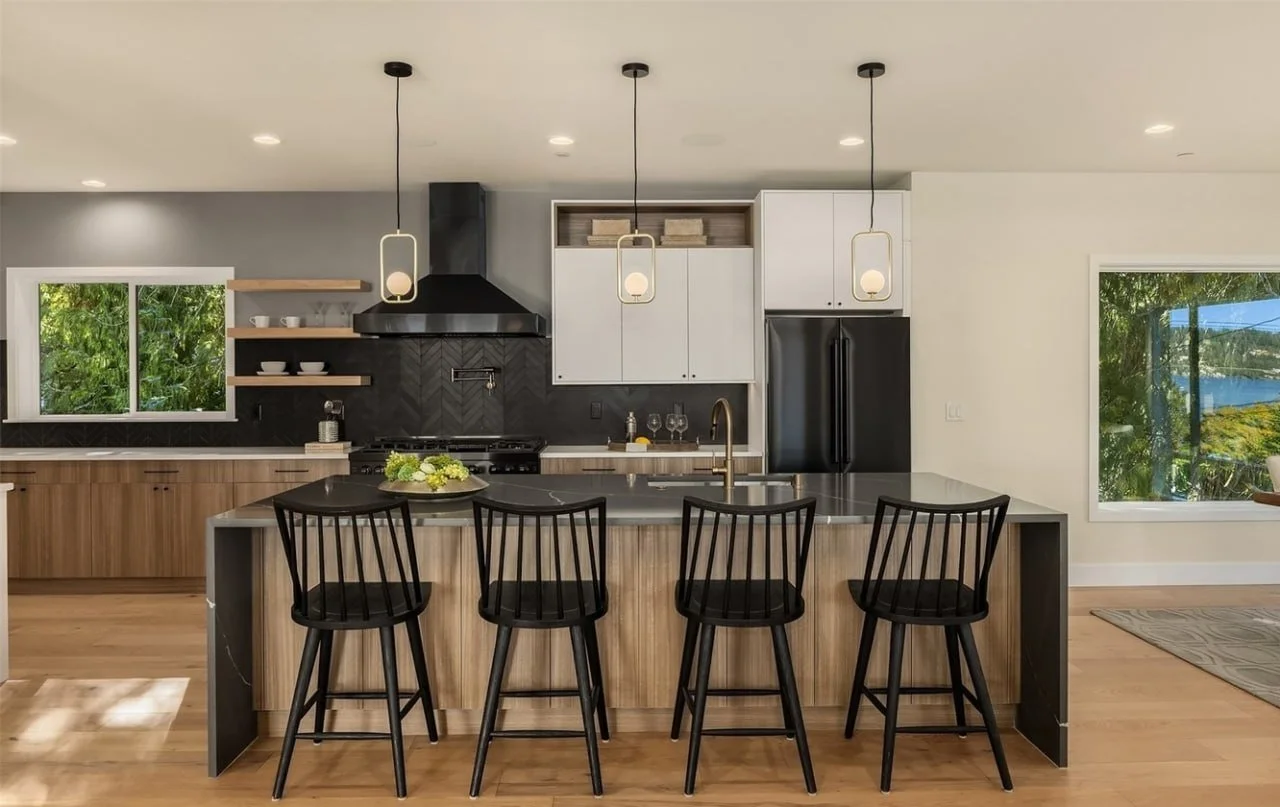 Modern kitchen with wooden cabinets, black appliances, white upper cabinets, open shelves, a black range hood, three pendant lights, black bar stools, window views of greenery and water.