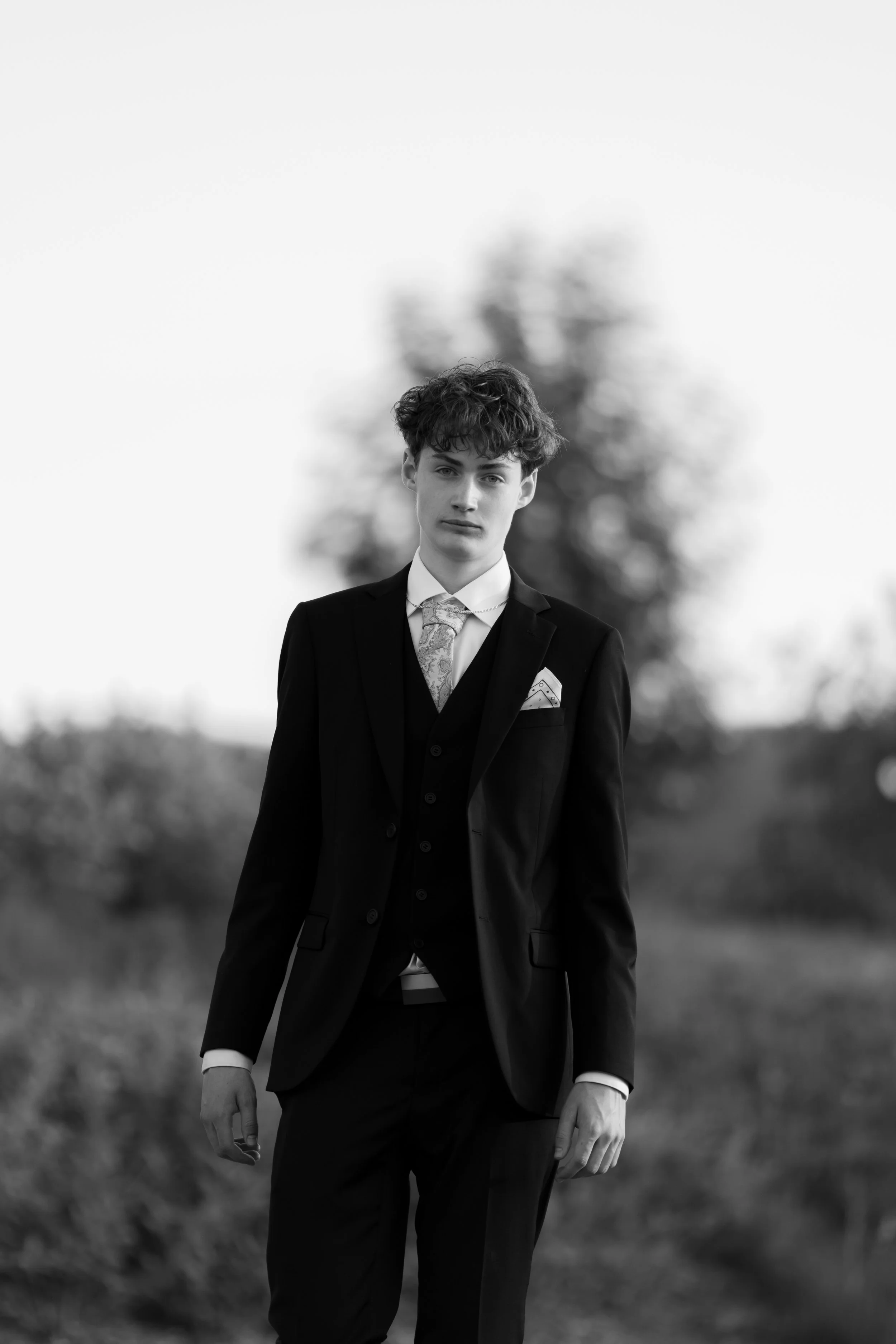 A young man in a dark suit and patterned tie, standing outdoors with a blurred natural background, in black and white.