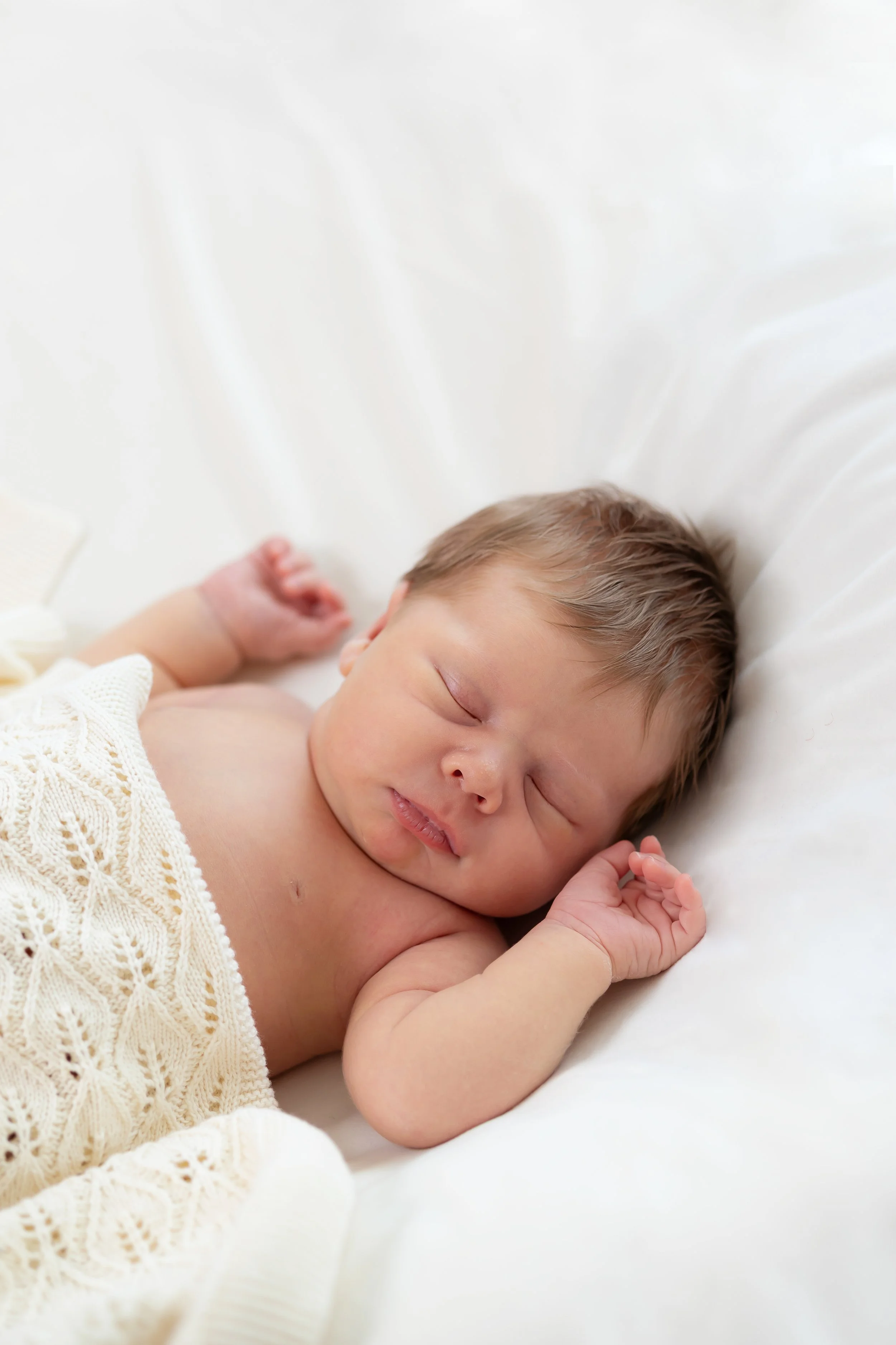 Close-up of a sleeping newborn baby lying on a white surface, wearing a cream-colored knitted blanket.