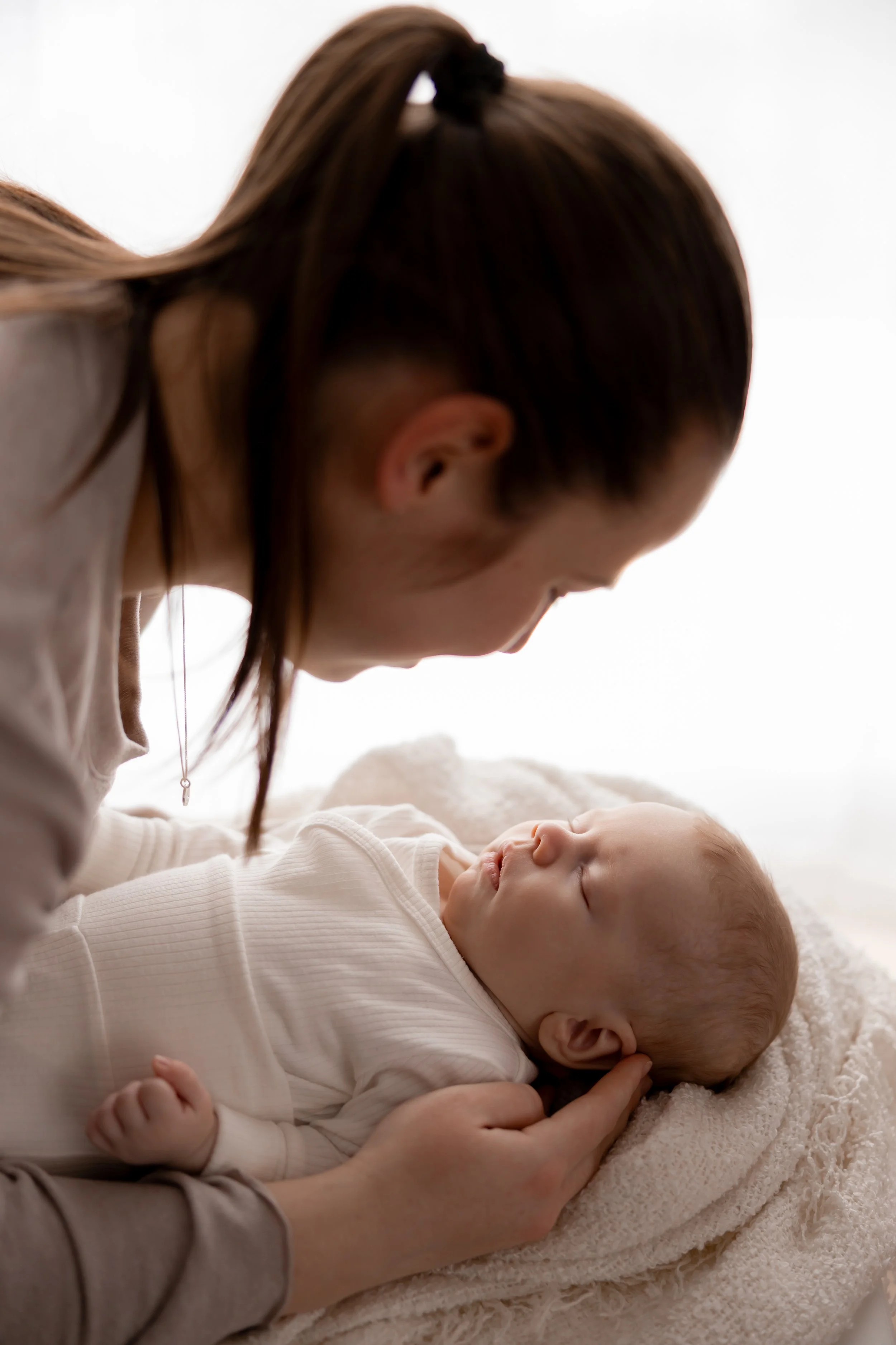 A woman gently holds a sleeping baby on a soft blanket, gazing at the baby in a tender moment.