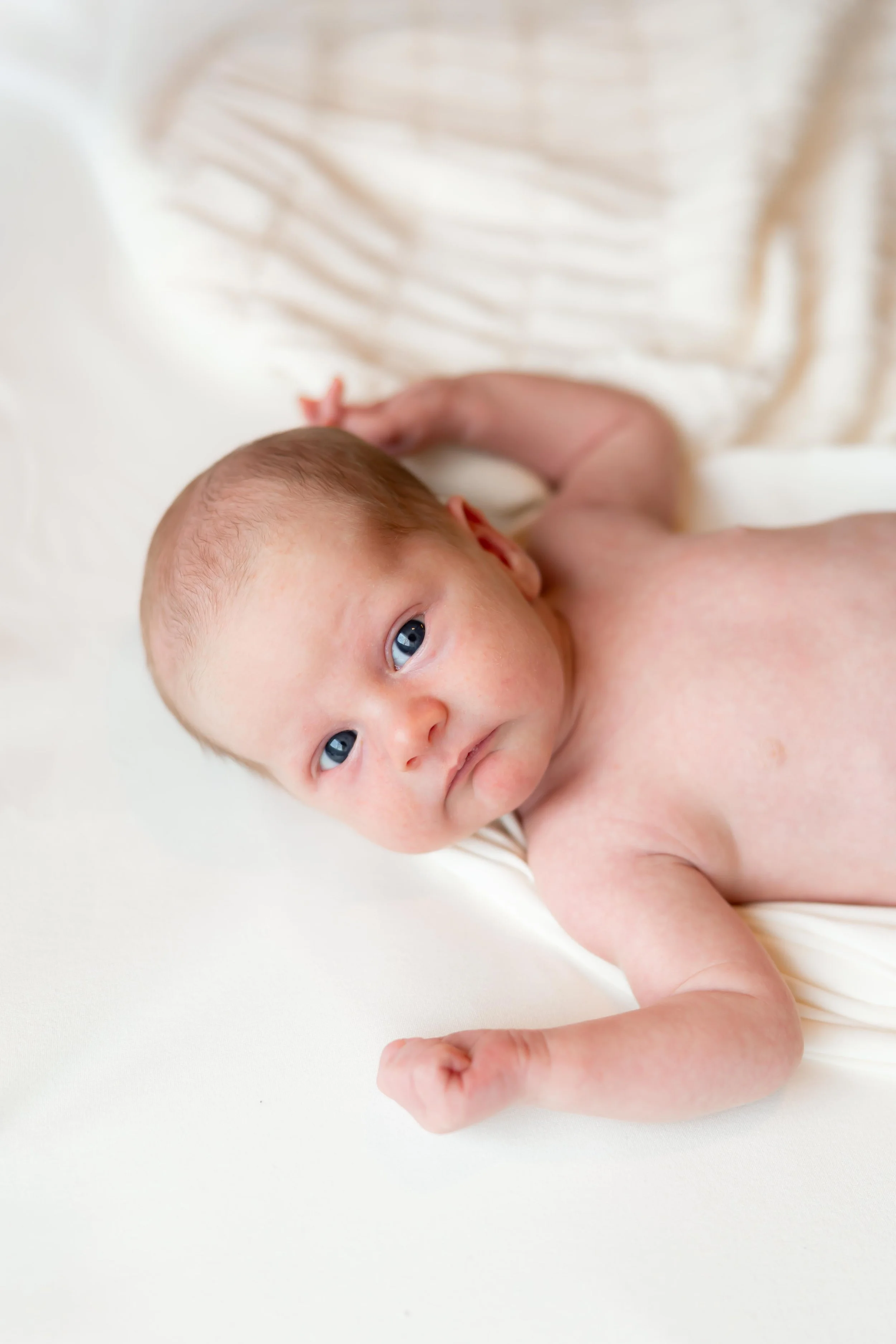 A newborn baby lying on a white surface, looking at the camera with blue eyes, with one arm bent and the other resting near the head.