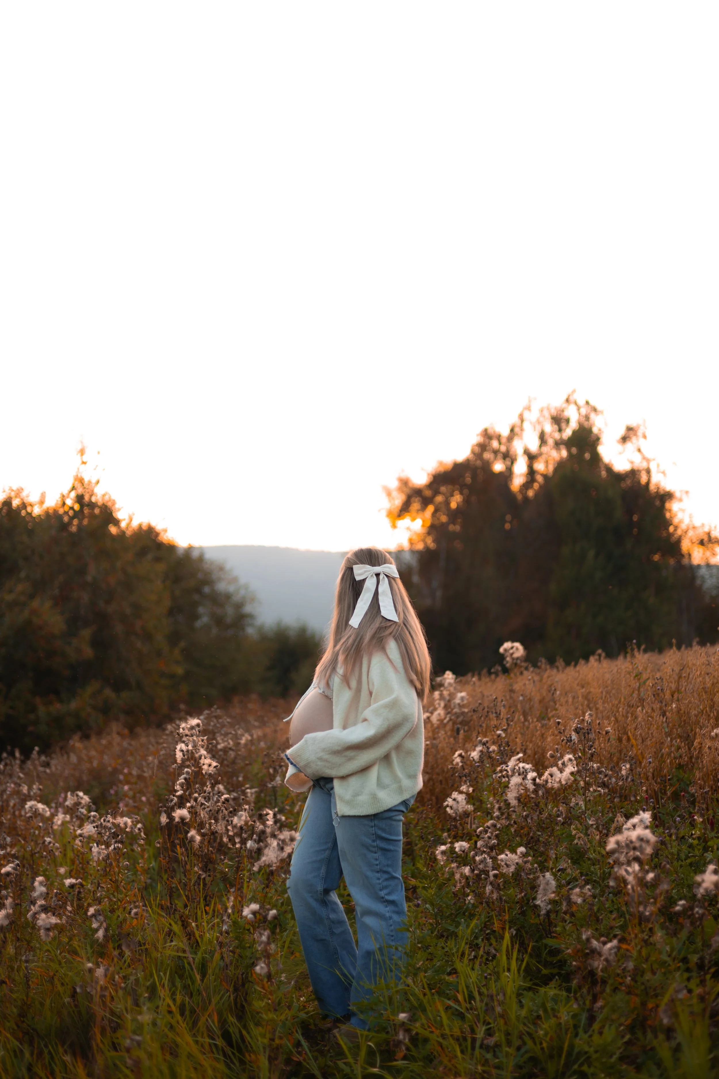 A pregnant woman standing in a field of wildflowers at sunset, looking away from the camera, with trees in the background.