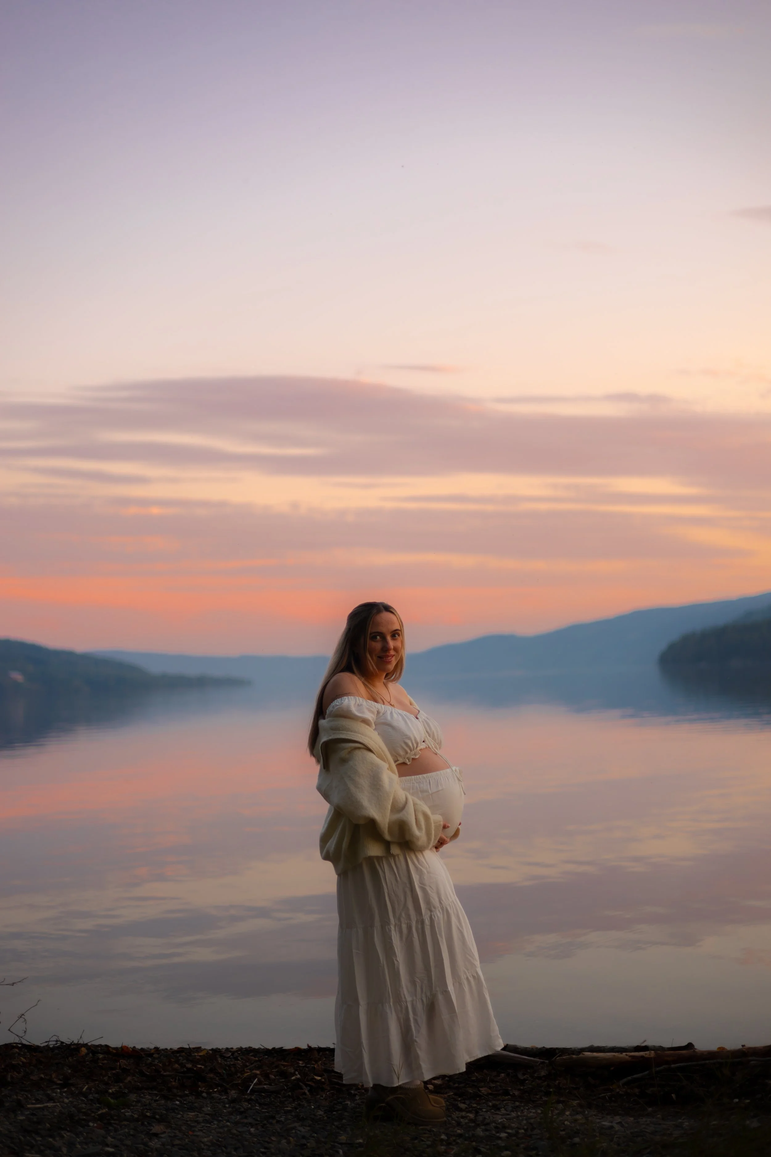 A pregnant woman standing by a lake at sunset, smiling and holding her belly, with mountains in the background.
