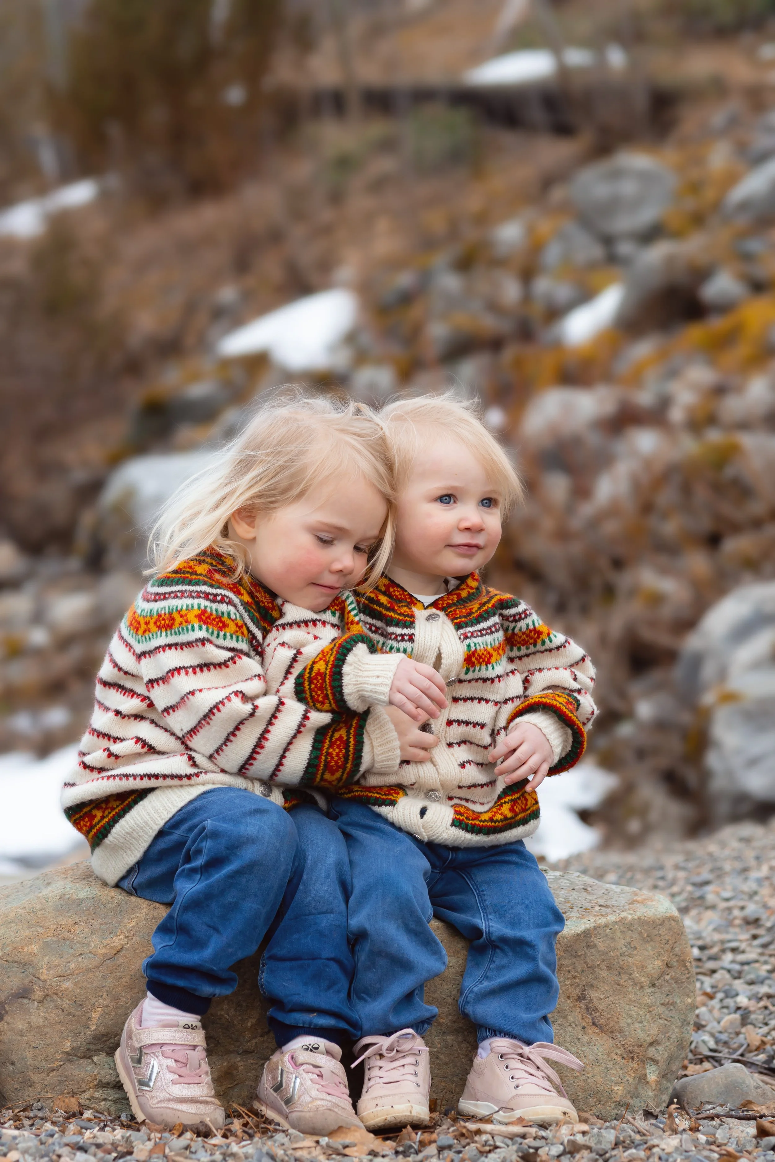 Two young girls sitting on a large rock outdoors during winter, wearing colorful sweaters and jeans, with a blurred background of rocks, trees, and patches of snow.
