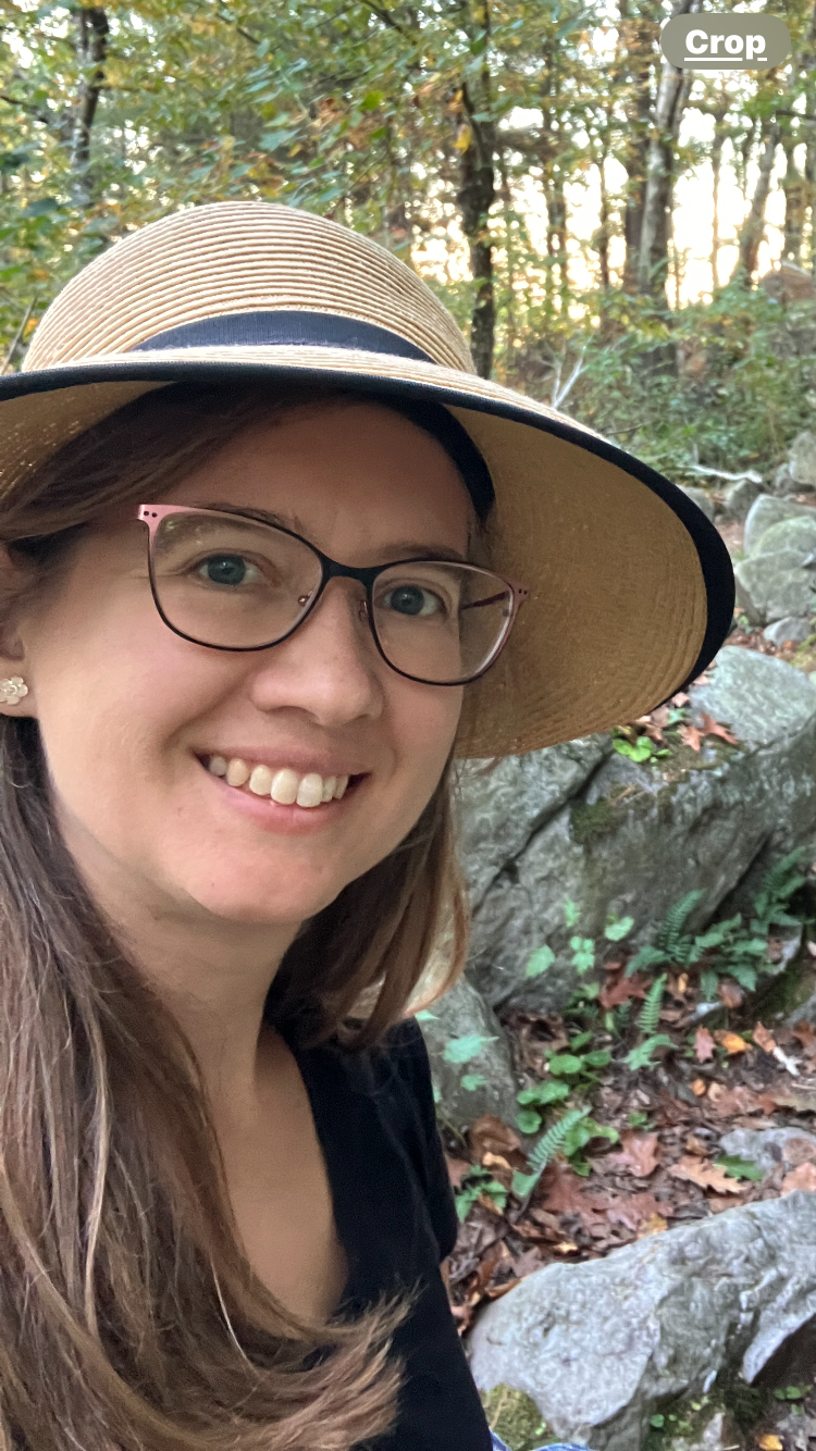 A woman smiling outdoors in a wooded area wearing a straw hat and glasses.