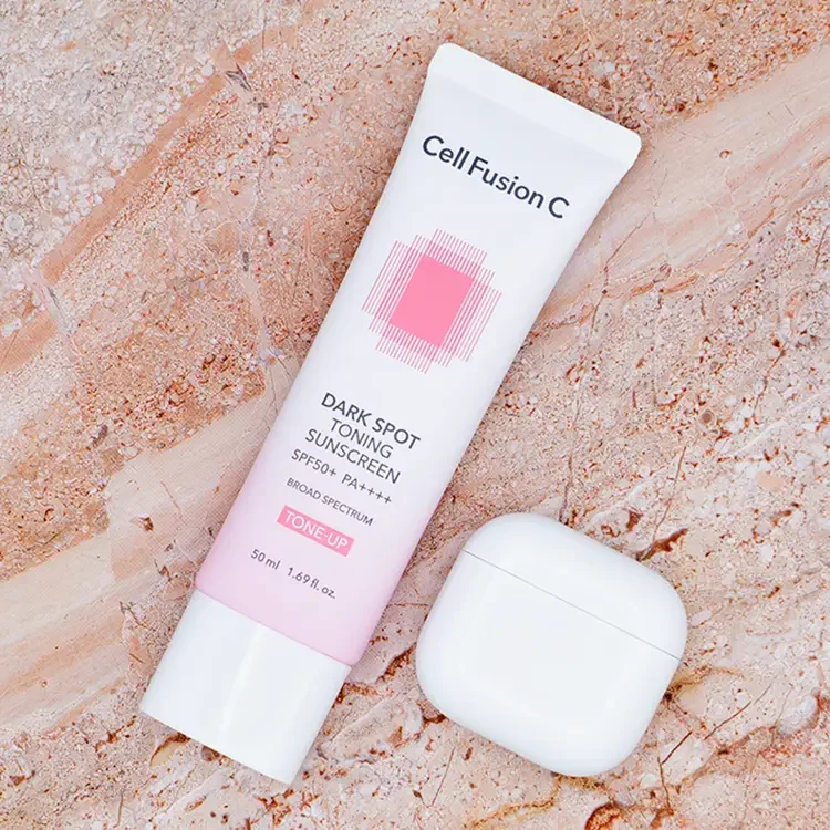 A square crop top-down view of Cell Fusion C Dark Spot Toning Sunscreen next to a white AirPods case on a luxury beige marble surface for size comparison, featured on TheWellnessExaminer.
