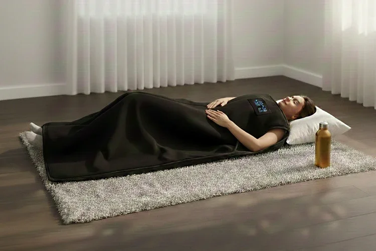 A person lying comfortably on a gray rug in a bright room, fully covered by a black infrared sauna blanket for full-body wellness.