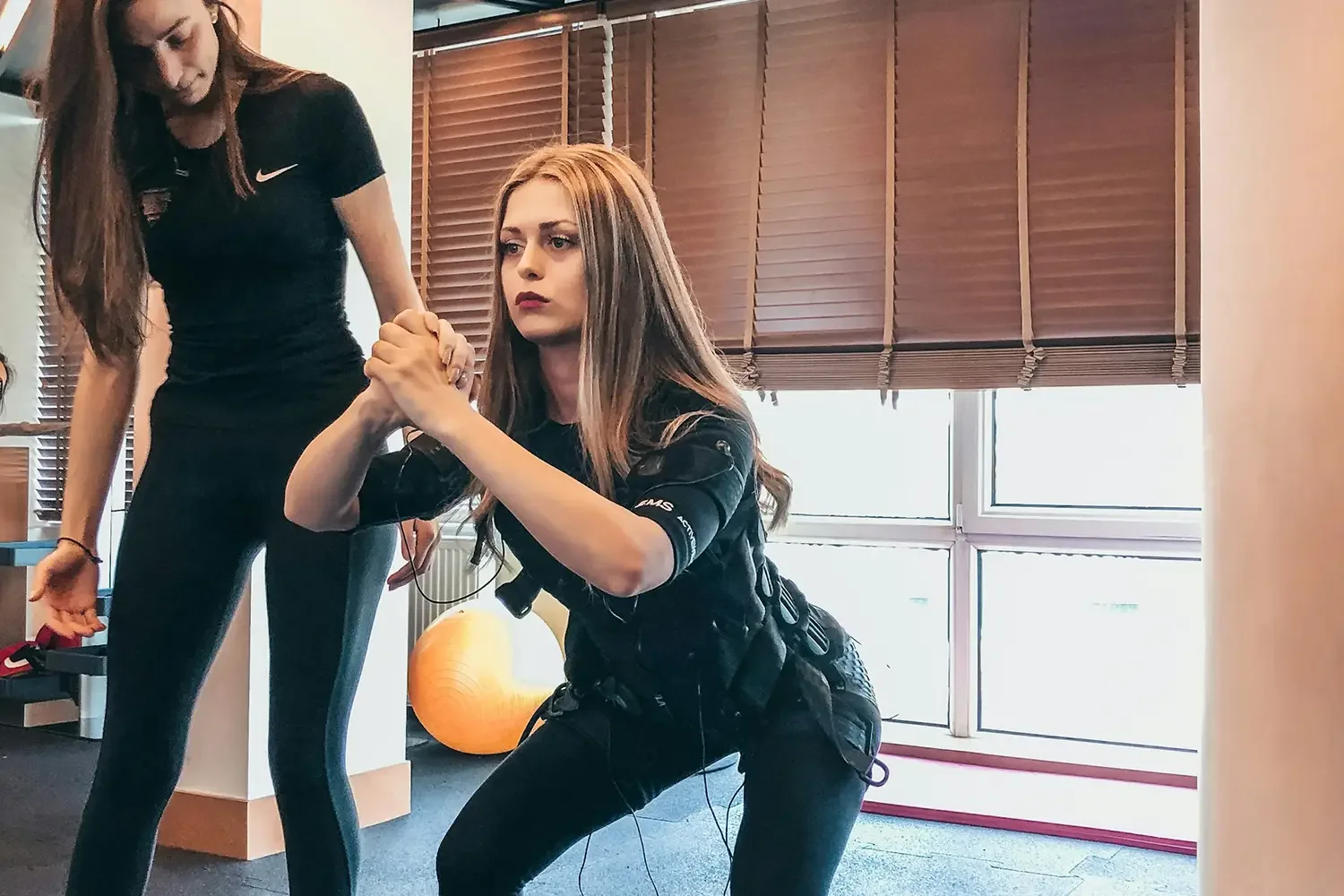A blonde woman in a gym setting performing a bodyweight squat while wearing a black whole-body EMS (Electrical Muscle Stimulation) training suit with adjustable straps, electrodes, and dangling connection wires.