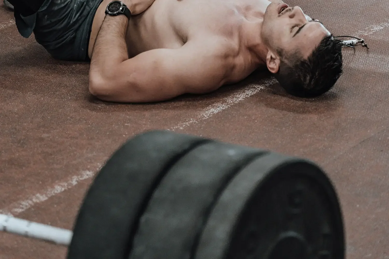 An exhausted young man lying flat on his back on a gym floor gasping for air next to a heavy barbell after a high intensity weightlifting workout.