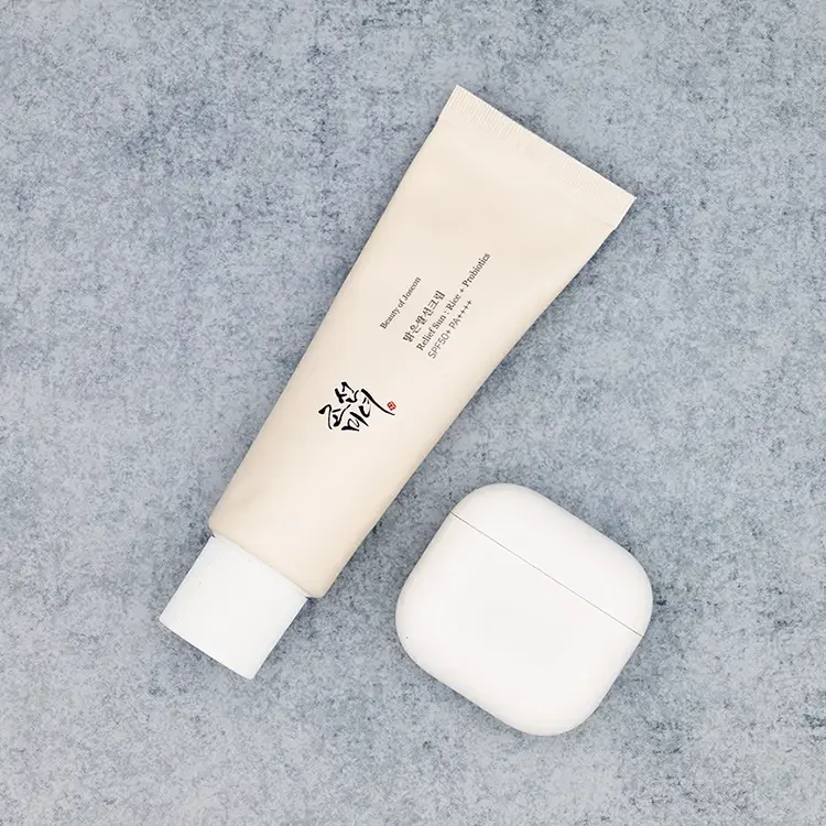 A square crop top-down view of Beauty of Joseon Relief Sun : Rice + Probiotics sunscreen tube next to a white AirPods case on a textured blue stone surface for size comparison, featured on TheWellnessExaminer.