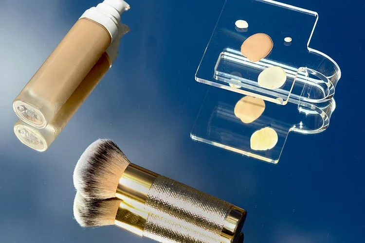 Liquid foundation bottle with a gold makeup brush and a clear acrylic mixing palette reflecting on a blue surface.