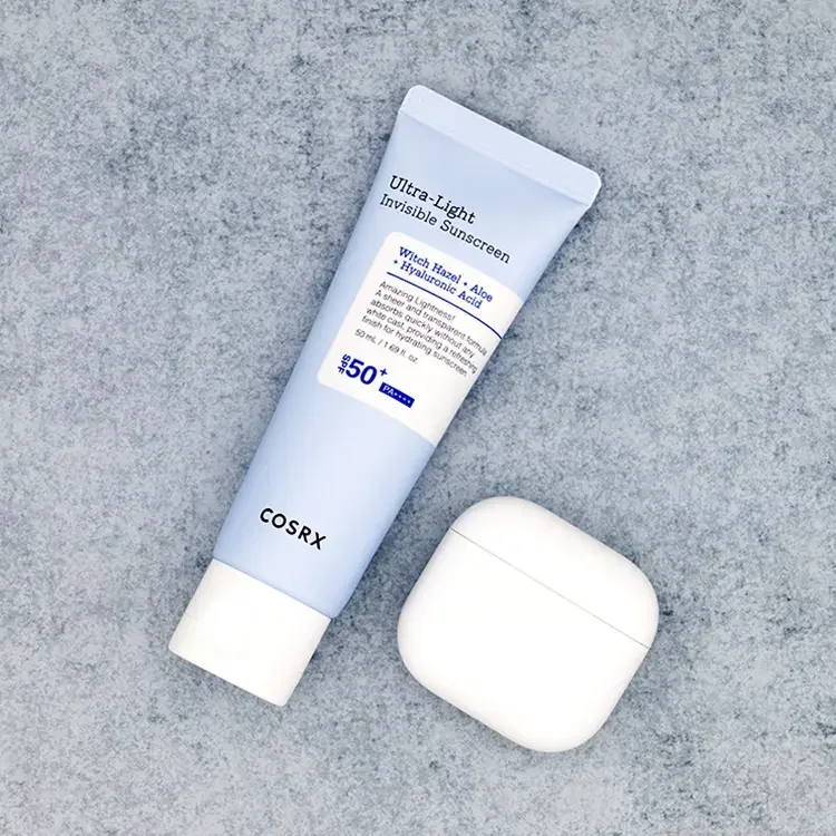 A square crop top-down view of COSRX Ultra-Light Invisible Sunscreen tube next to a white AirPods case on a textured blue stone surface for size comparison, featured on TheWellnessExaminer.